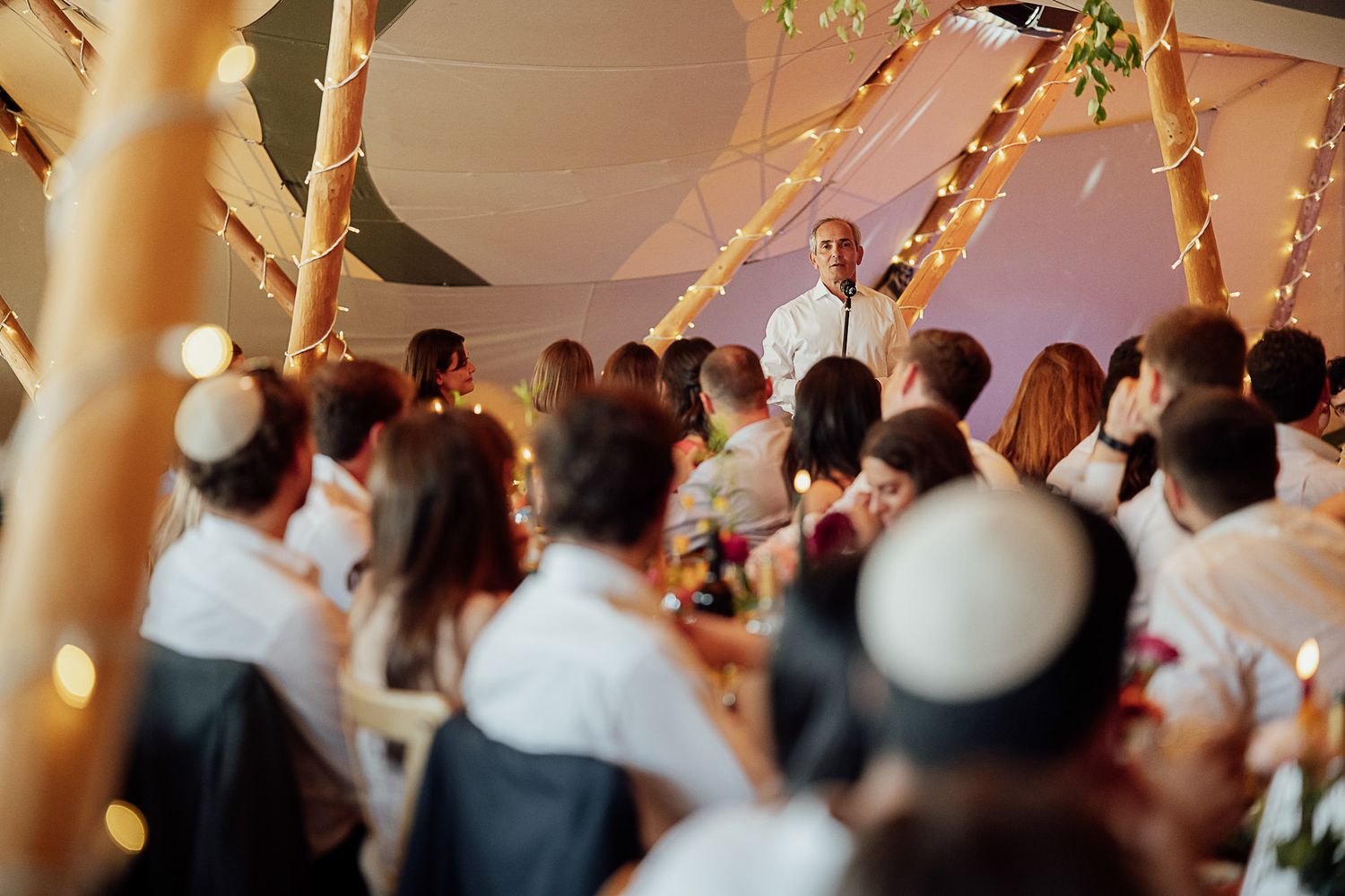 Guests seated under a decorated tent with fairy lights listen to a speaker at an elegant outdoor wedding reception.