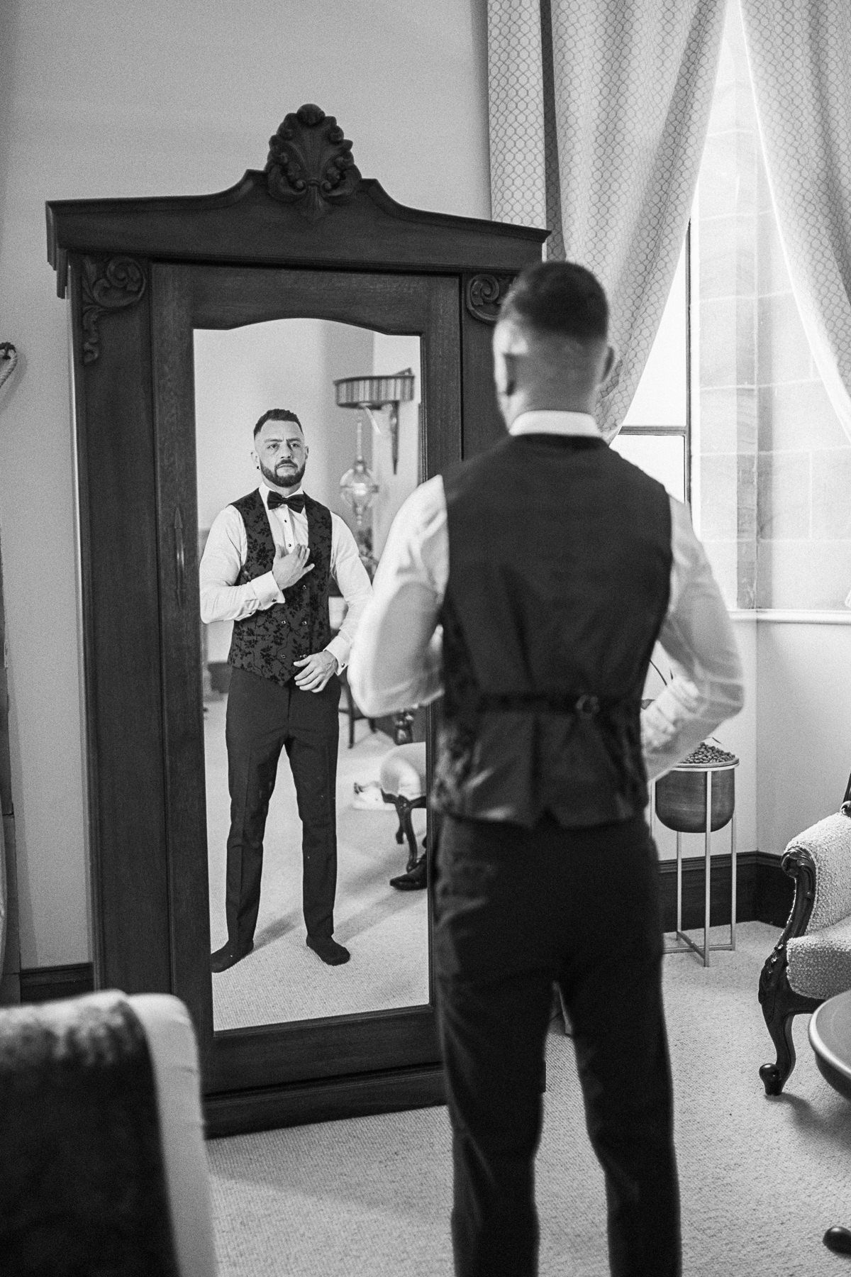 Groom in vest and bow tie checking his appearance in an ornate full-length mirror before wedding ceremony.