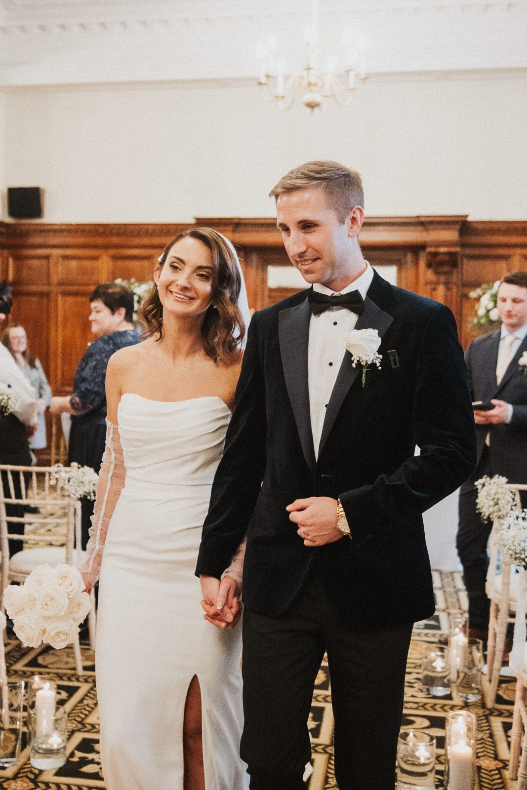 Newlyweds walking down aisle together past candle-lit decor in elegant reception venue.