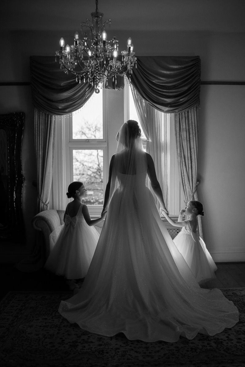 Black and white silhouette of wedding gowns displayed in front of sunlit windows with elegant drapes and chandeliers at West Tower venue.