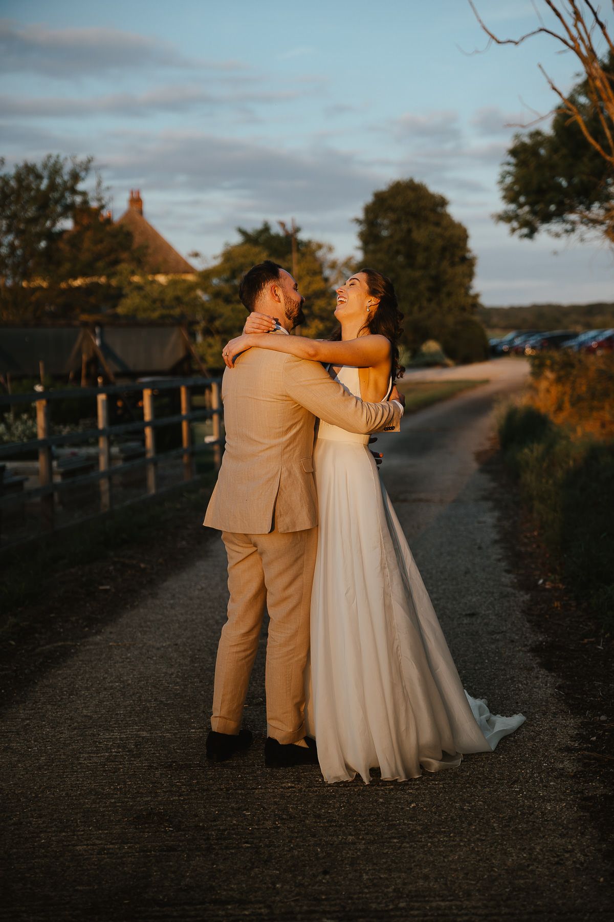 A couple embraces on a dirt path at sunset wearing beige suit and white dress as the golden light illuminates them.