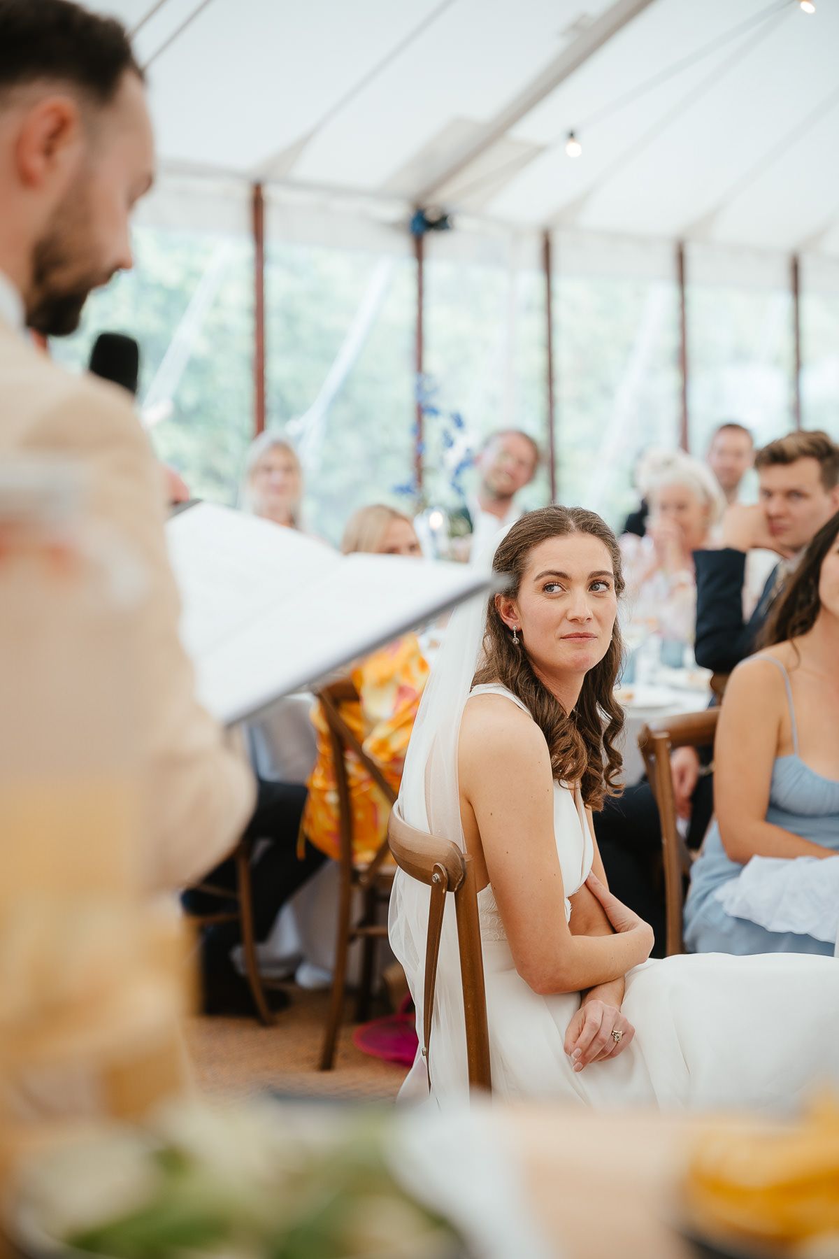 Wedding reception in a bright tent venue with guests seated at tables listening to speeches.