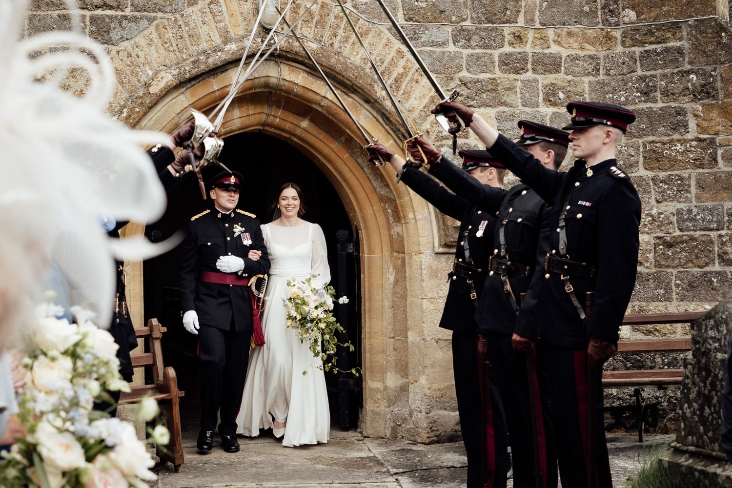 Military officers in formal dress uniform create traditional saber arch outside stone church archway during wedding ceremony.