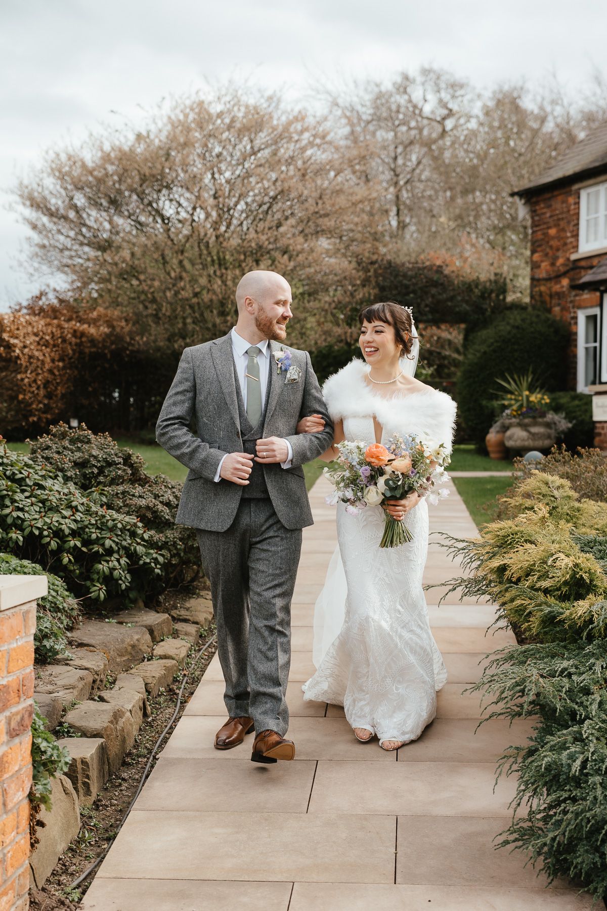 A wedding couple walking along a garden path on their special day in autumn, with her carrying a peach floral bouquet.