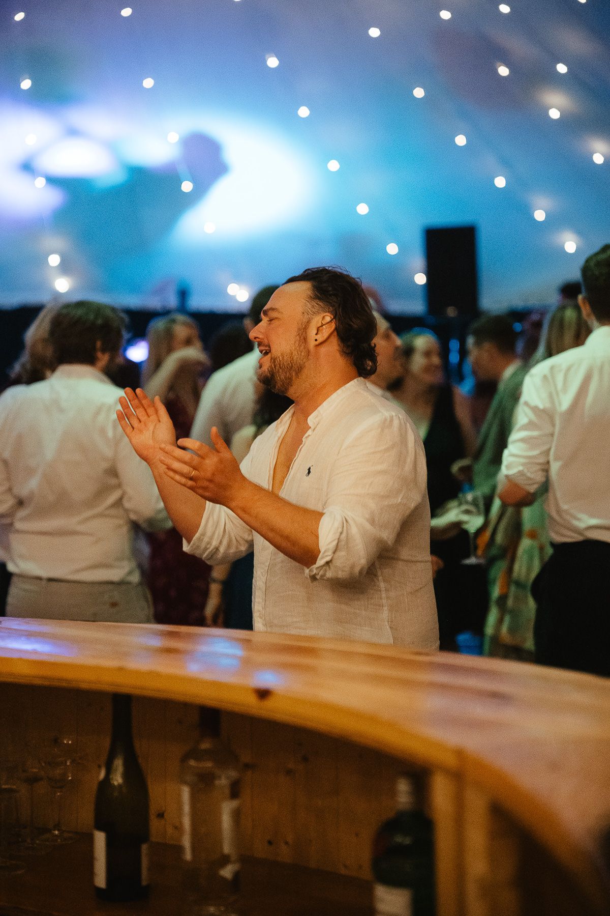 A person clapping and laughing joyfully at a festive event with twinkling lights and a curved bar in the foreground.