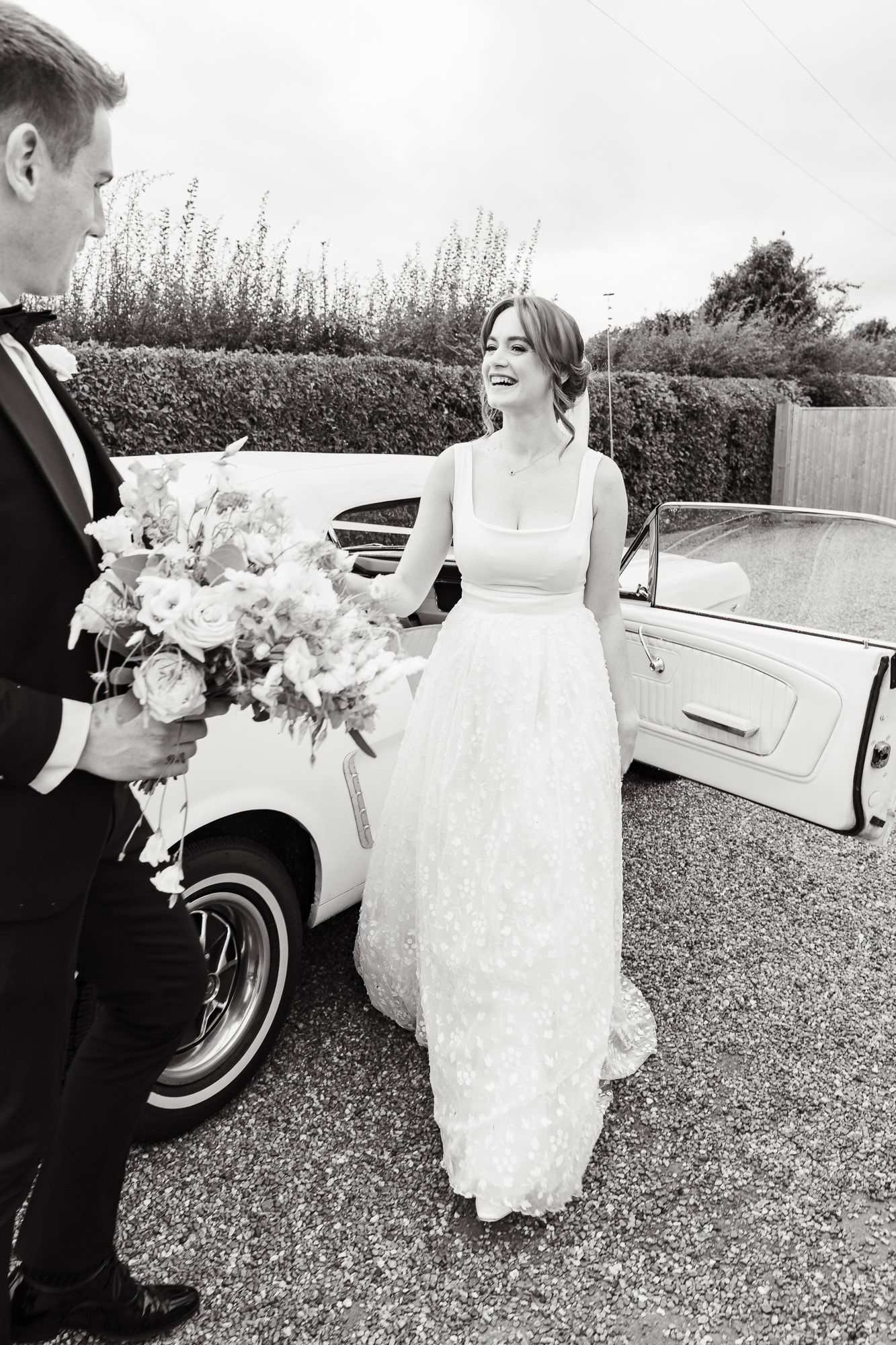 A romantic moment between newlyweds by a classic car as the groom presents a beautiful floral bouquet.