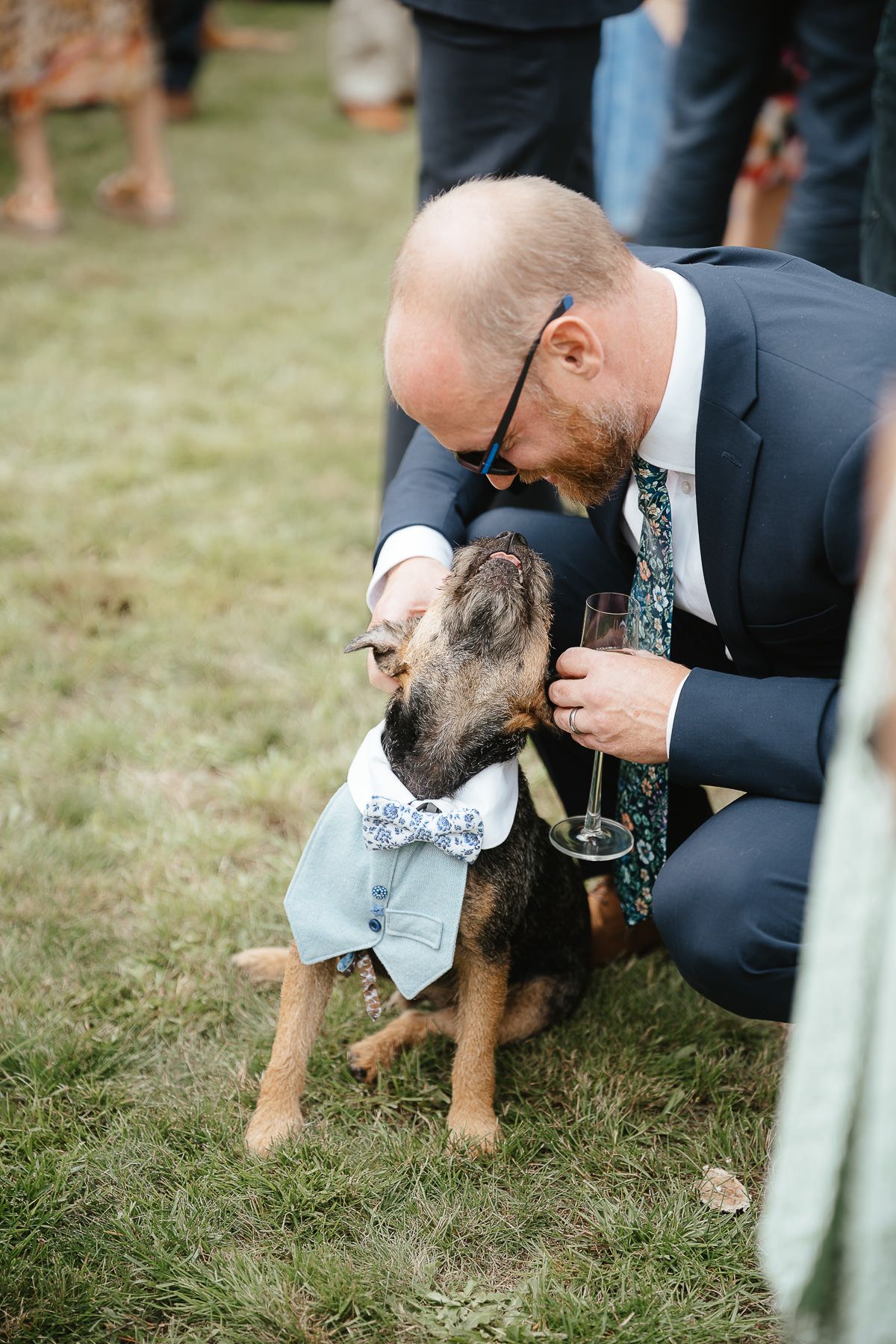A dog wearing a bow tie and dress shirt is lovingly embraced by someone in a navy suit at an outdoor event.