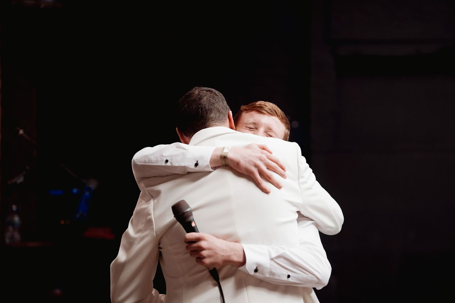 Two people in white formal attire share an emotional embrace during what appears to be a special event.