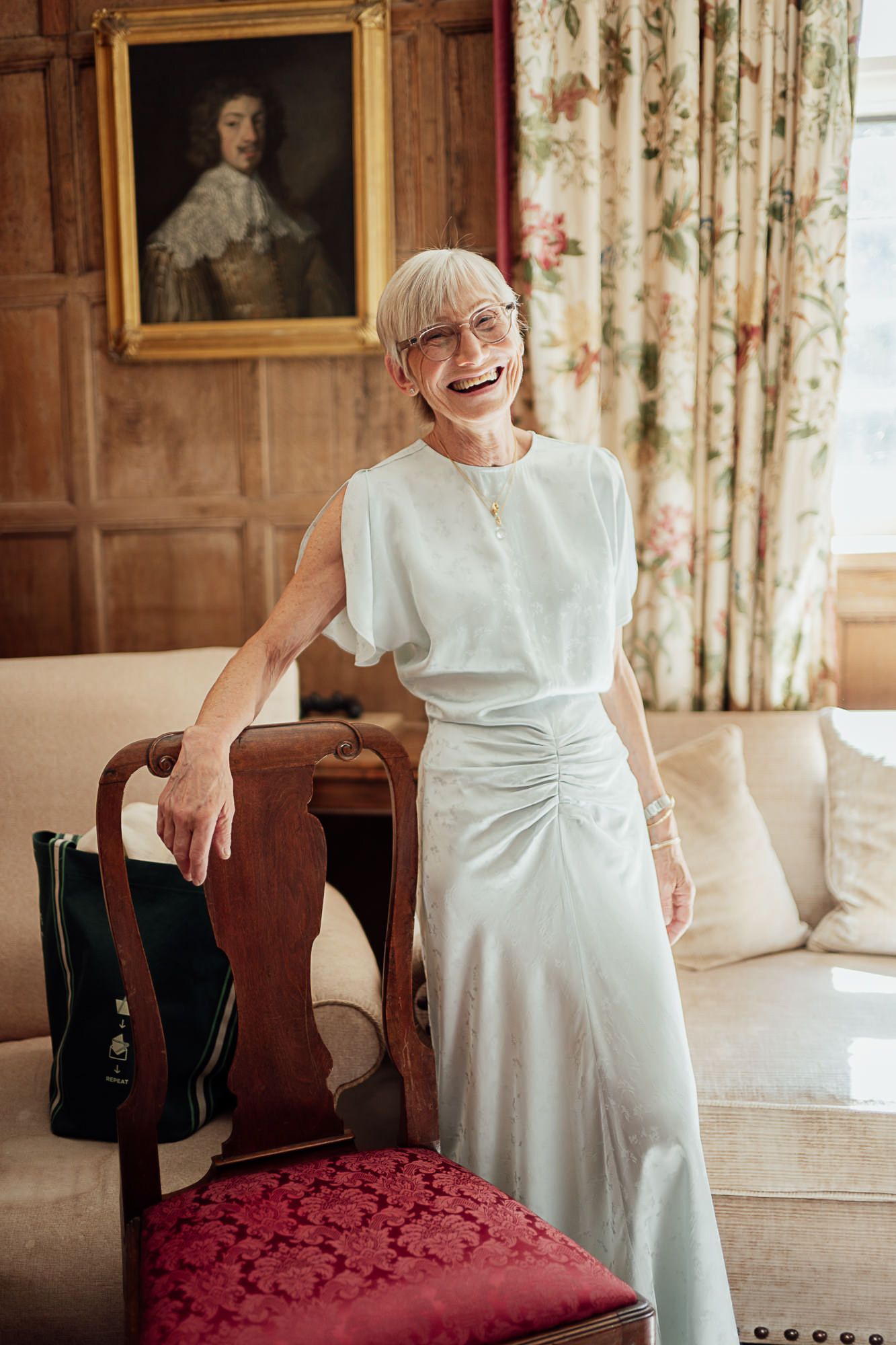 Elegant older woman in white satin dress laughing in a wood-paneled room with floral curtains and portrait painting.