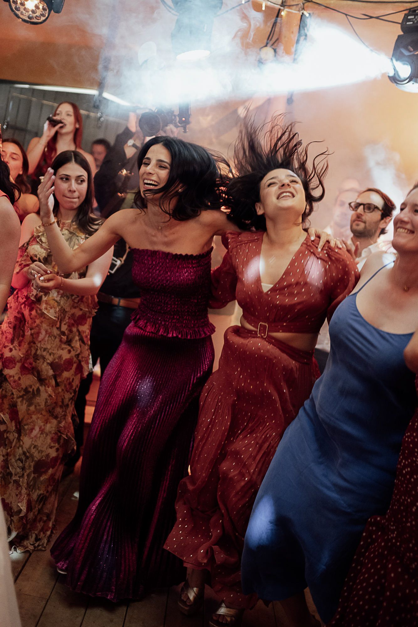 Two women in sparkling red dresses dancing joyfully at a lively party with guests and smoke effects.