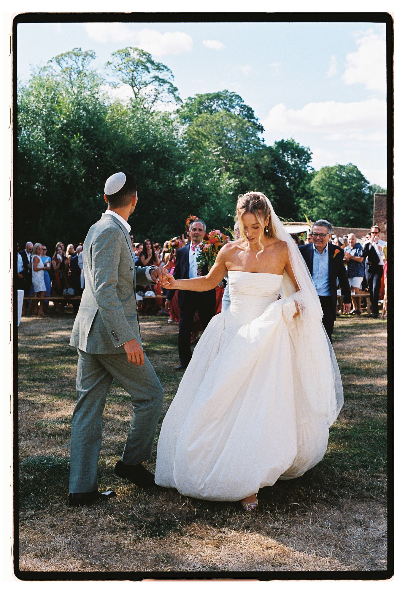 Bride in white gown and groom in gray suit dancing outdoors at a sunny garden wedding ceremony.
