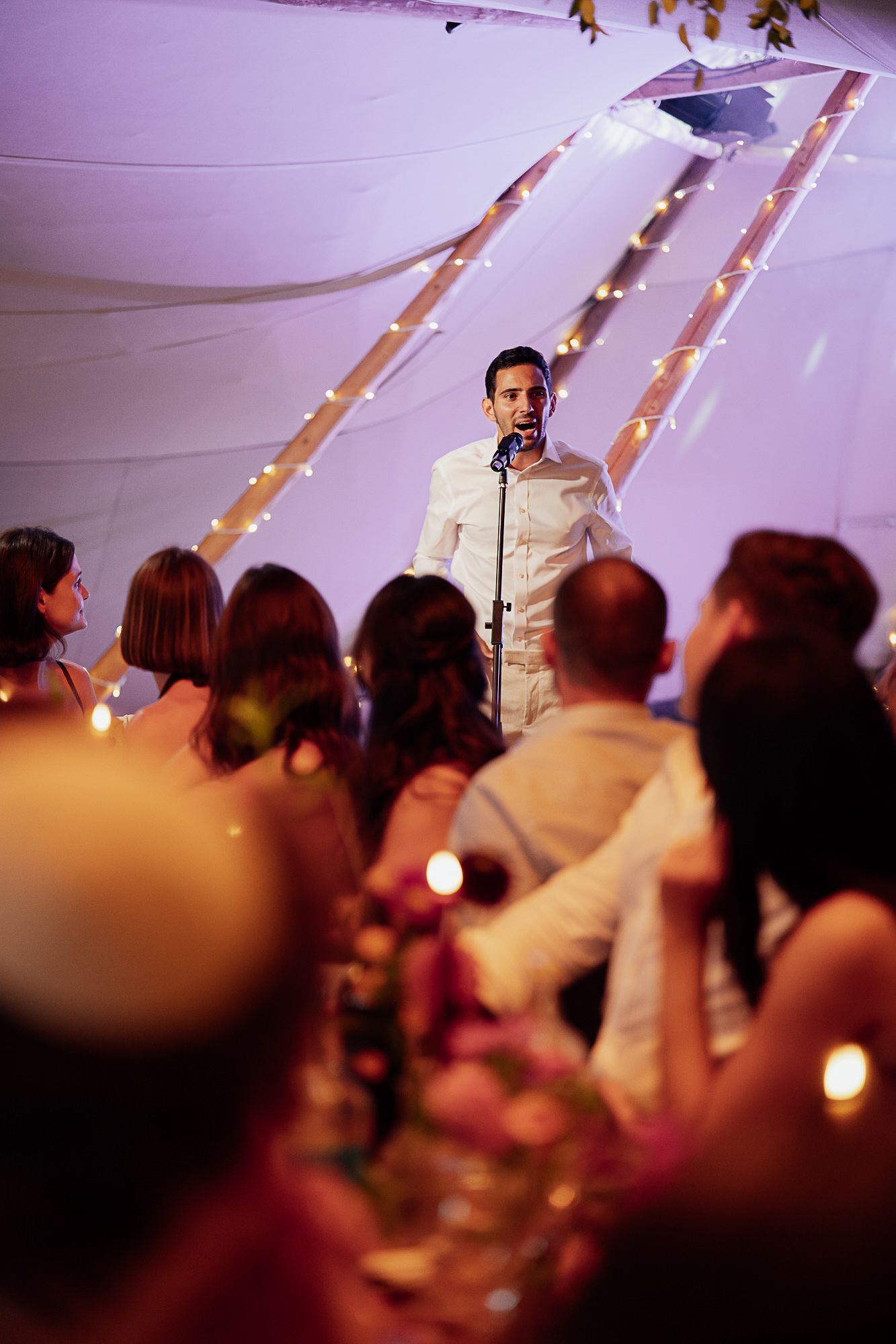 A man in white speaks at a microphone before seated wedding guests under a tent adorned with fairy lights.