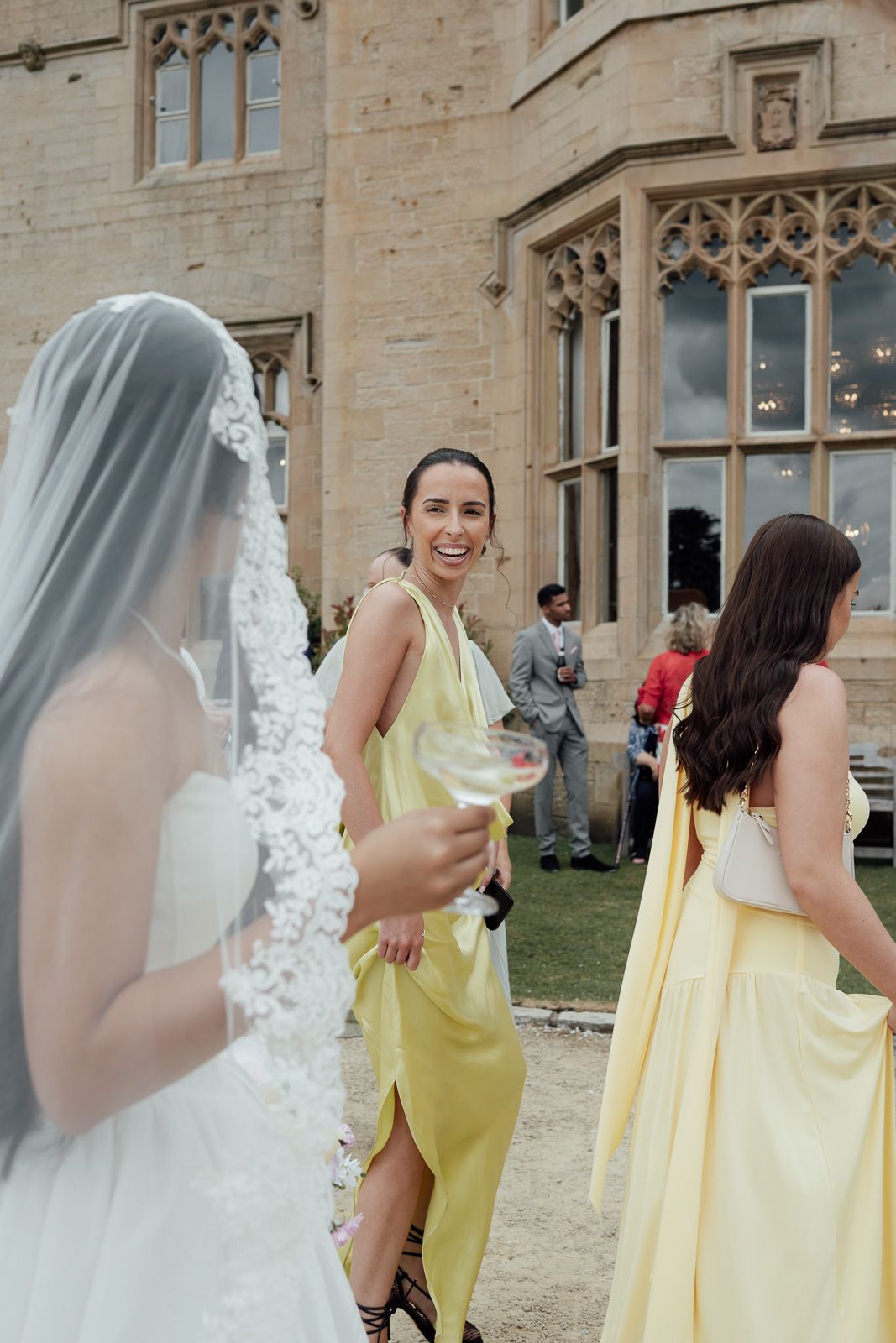 Bridesmaid in yellow dress laughs with bride in lace veil outside a grand stone manor.