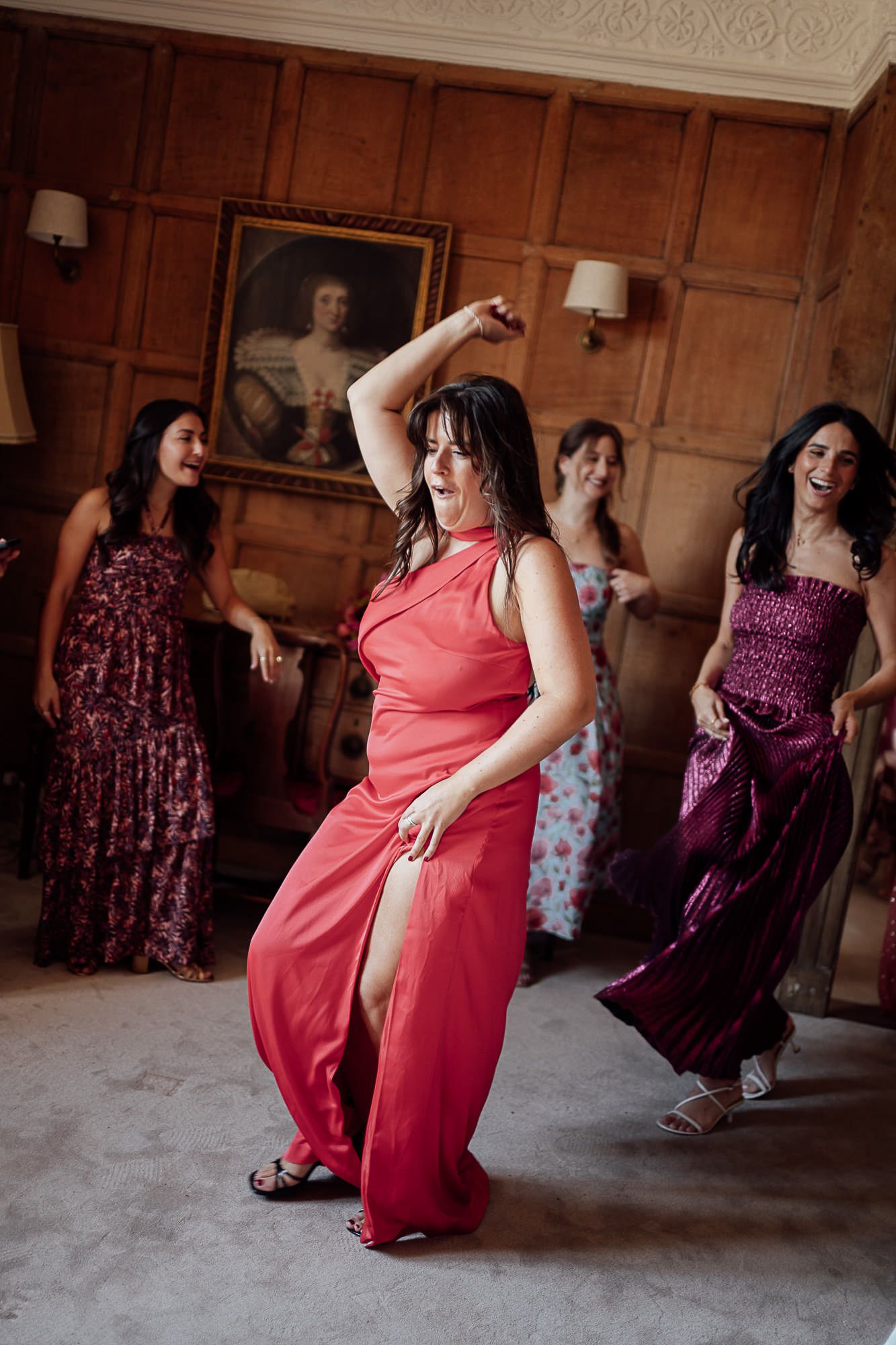 Women in elegant dresses dancing joyfully in a wood-paneled room with a portrait painting on the wall.