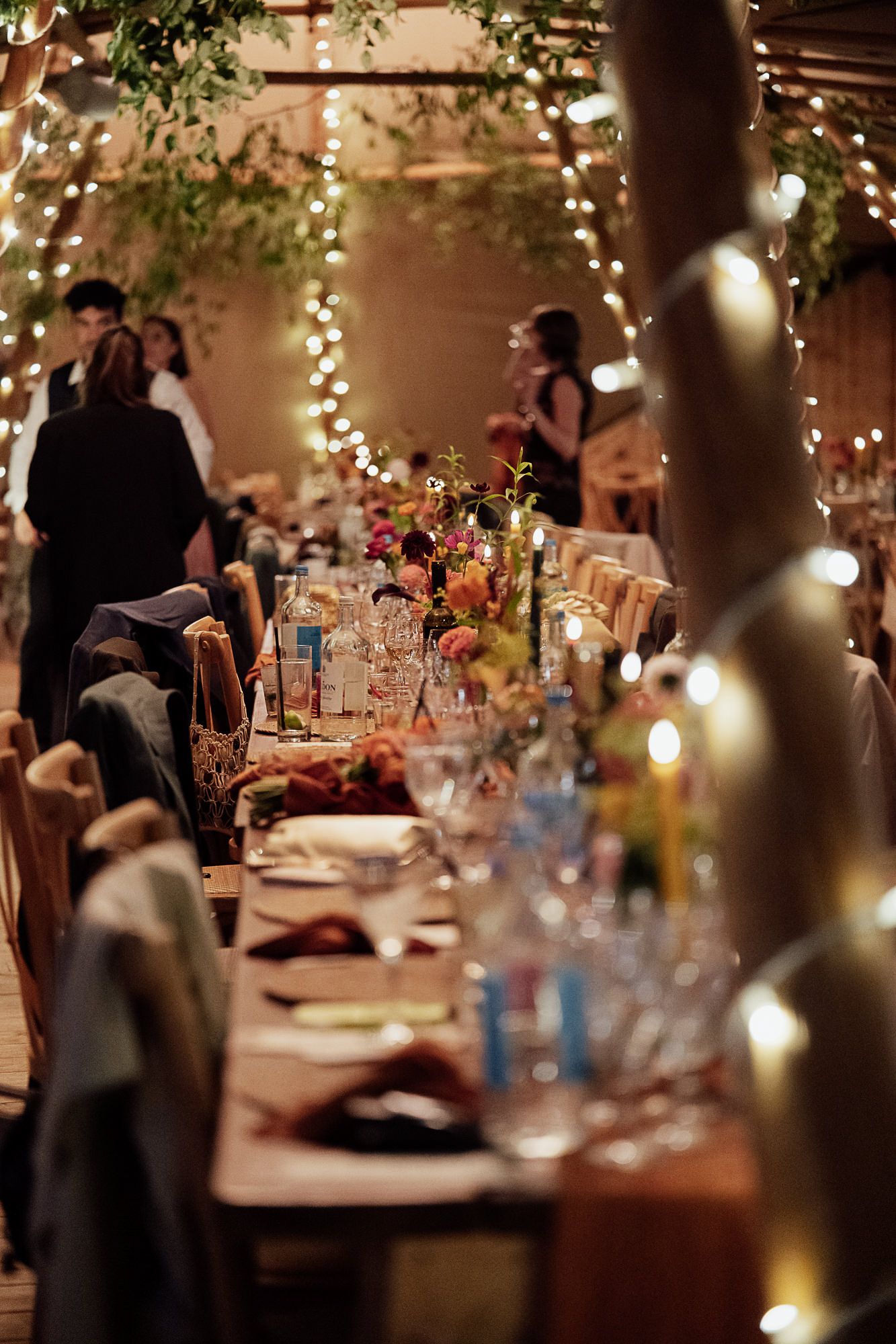 Elegant wedding reception with long dining table, fairy lights, floral centerpieces, and guests mingling in a warmly lit venue.