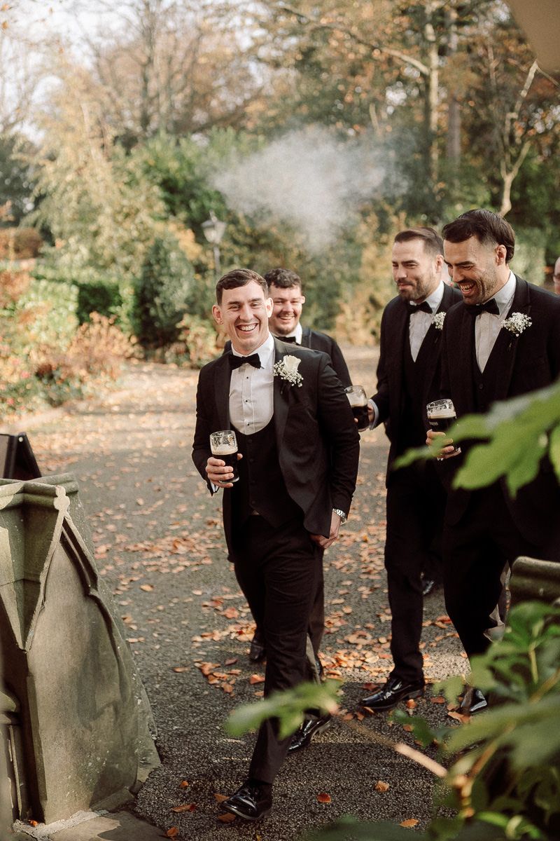 Groomsmen in black tuxedos laughing together on autumn grounds.
