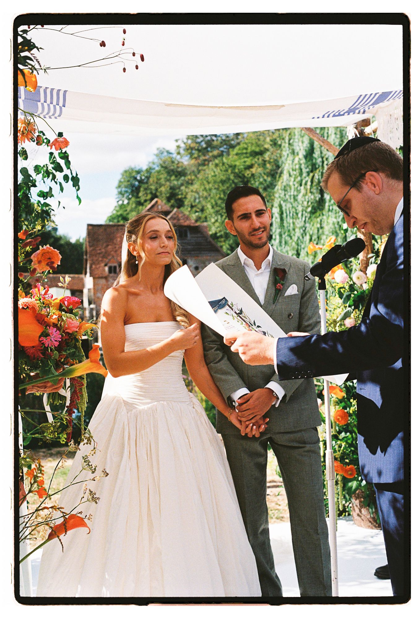 Bride and groom stand at outdoor wedding altar with colorful floral arrangements while officiant reads from a document.