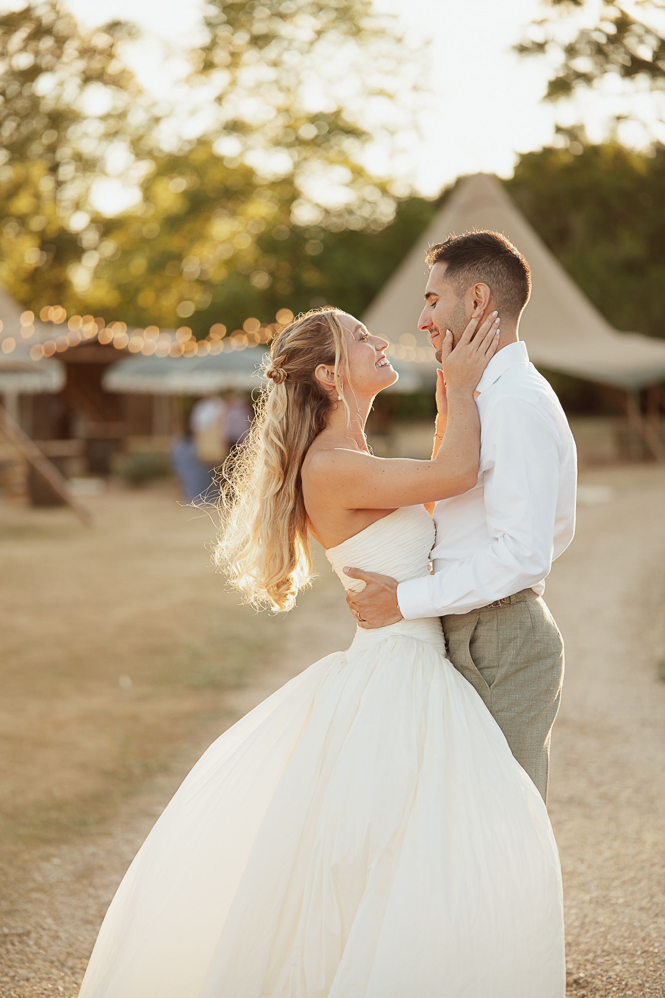 A bride and groom share a romantic dance outdoors at golden hour, with string lights and a rustic barn in the background.