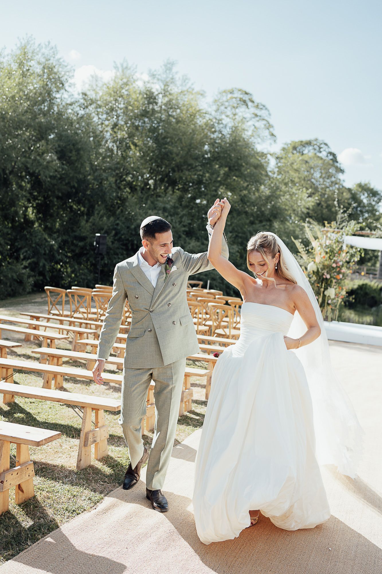 Bride and groom walking down outdoor aisle, holding hands raised, surrounded by wooden chairs and lush green trees.