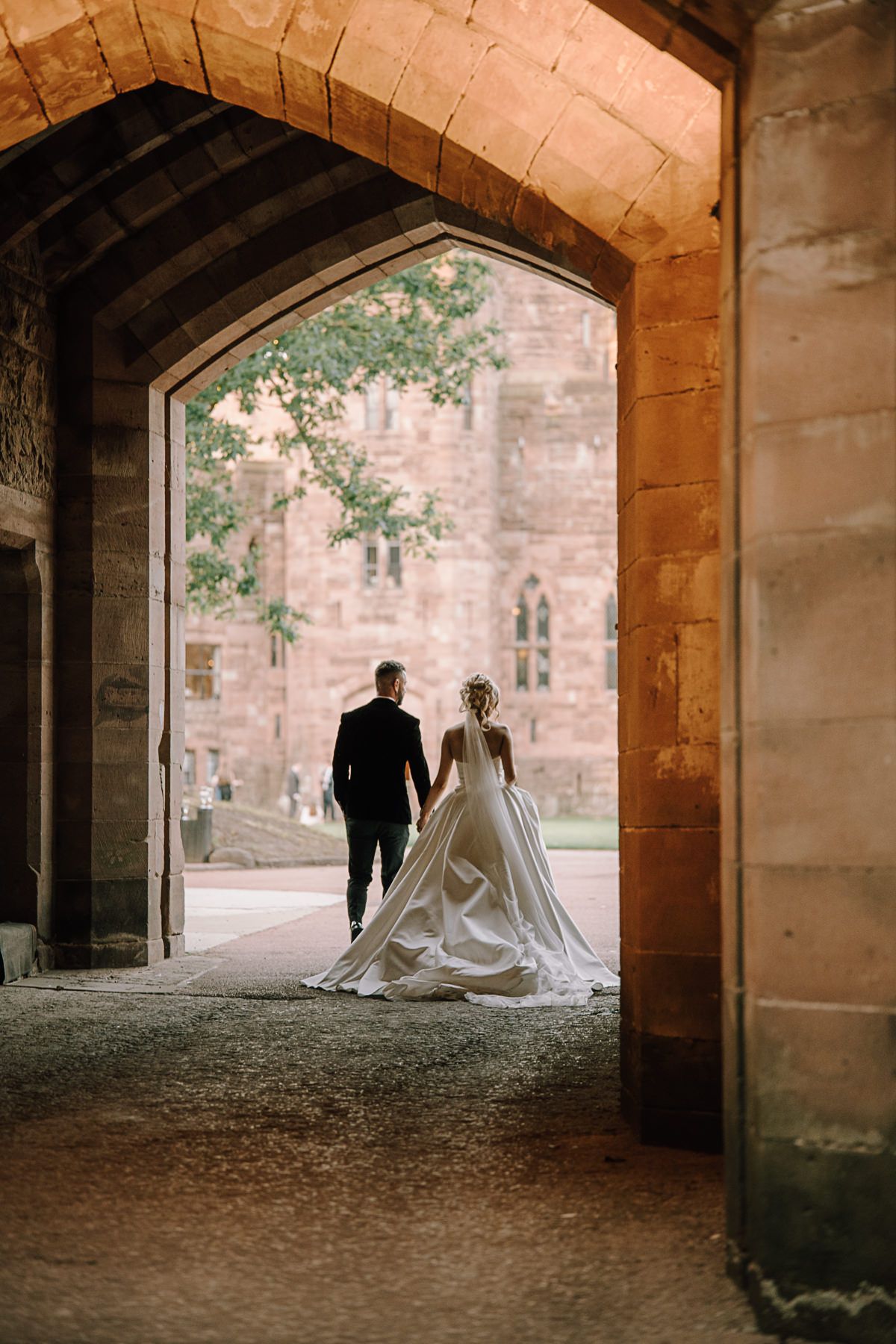 Bride and groom walking hand in hand through a stone archway toward a historic castle venue on their wedding day.
