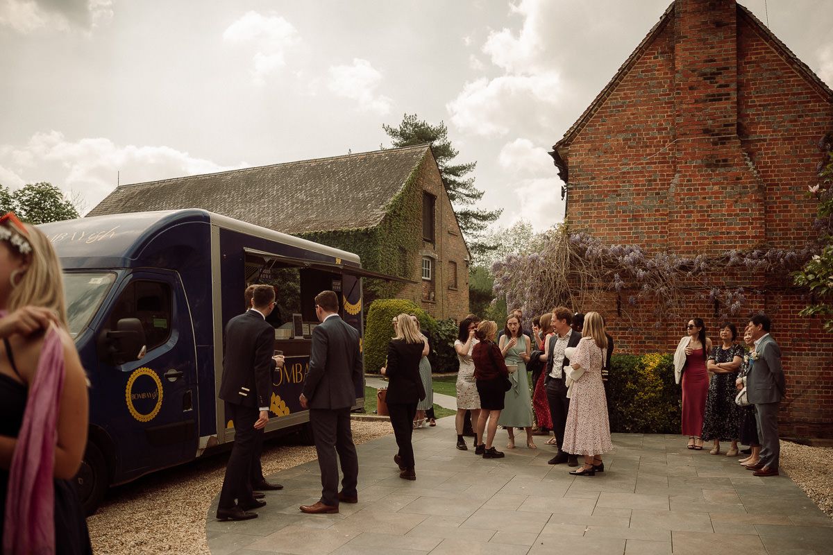 Wedding guests gather outside rustic brick barns during an outdoor countryside celebration.