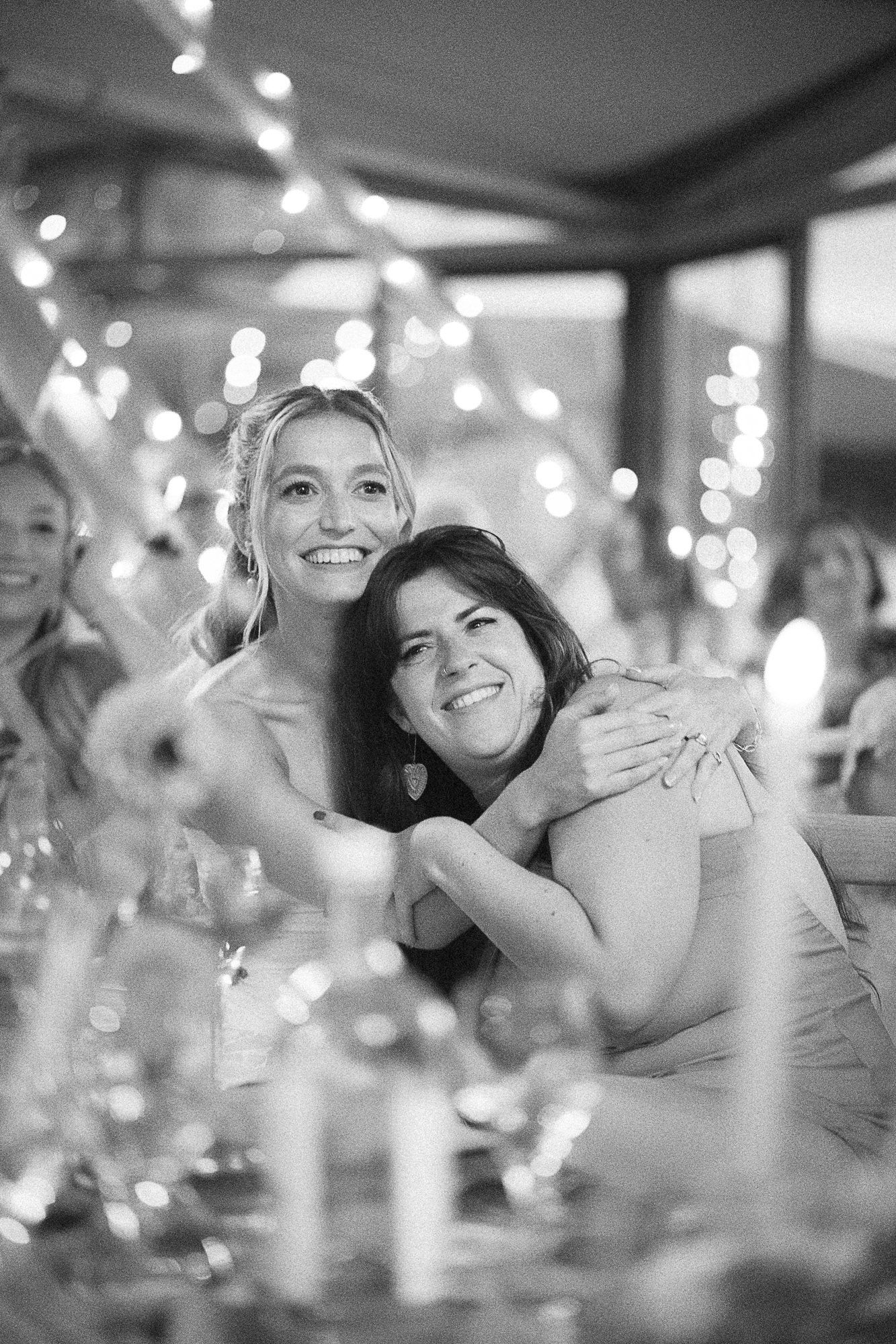 Two bridesmaids smiling and hugging at a wedding reception with bokeh string lights in black and white.