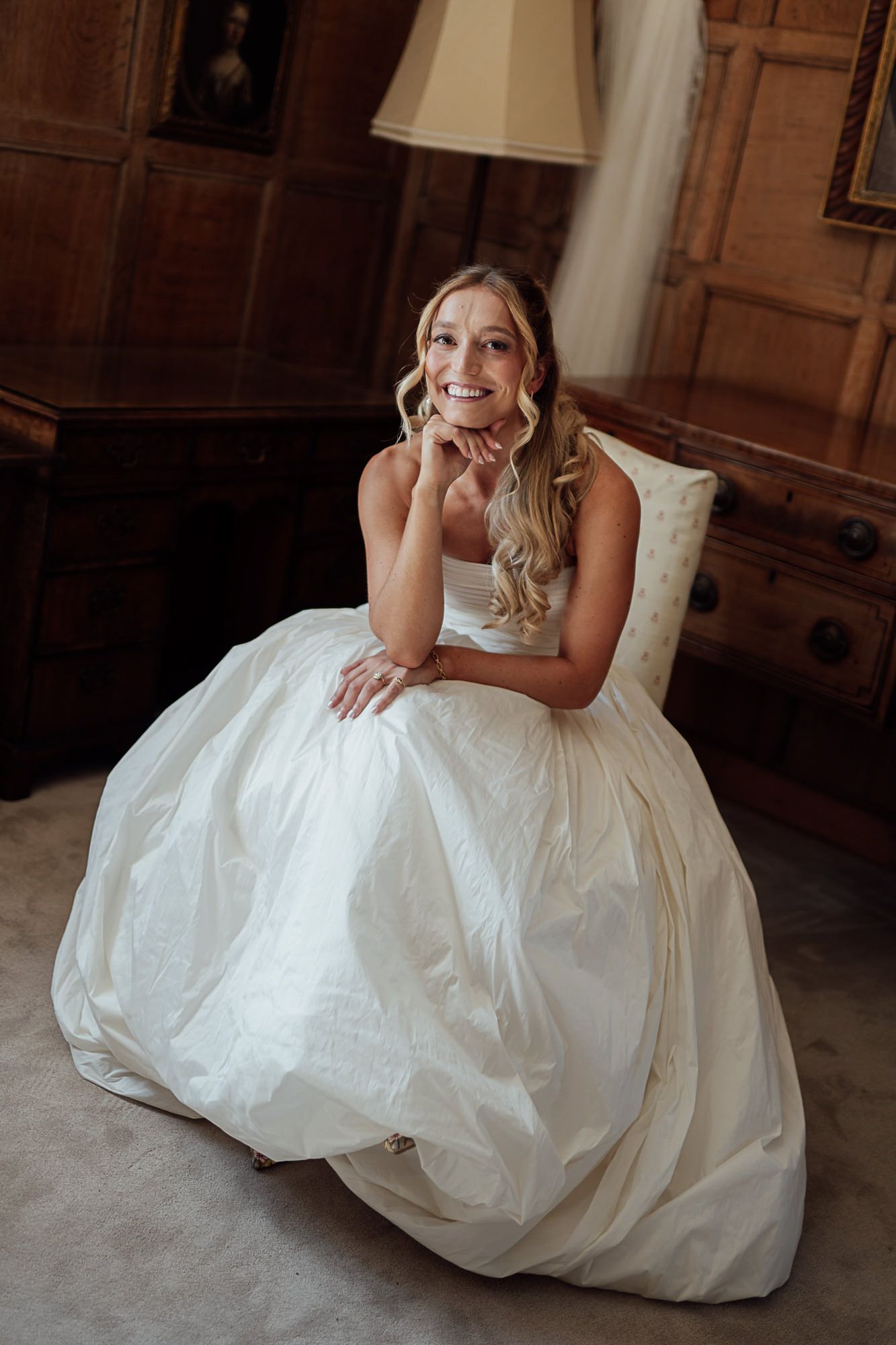 Smiling bride in a white ball gown sitting elegantly in a wood-paneled room on her wedding day.