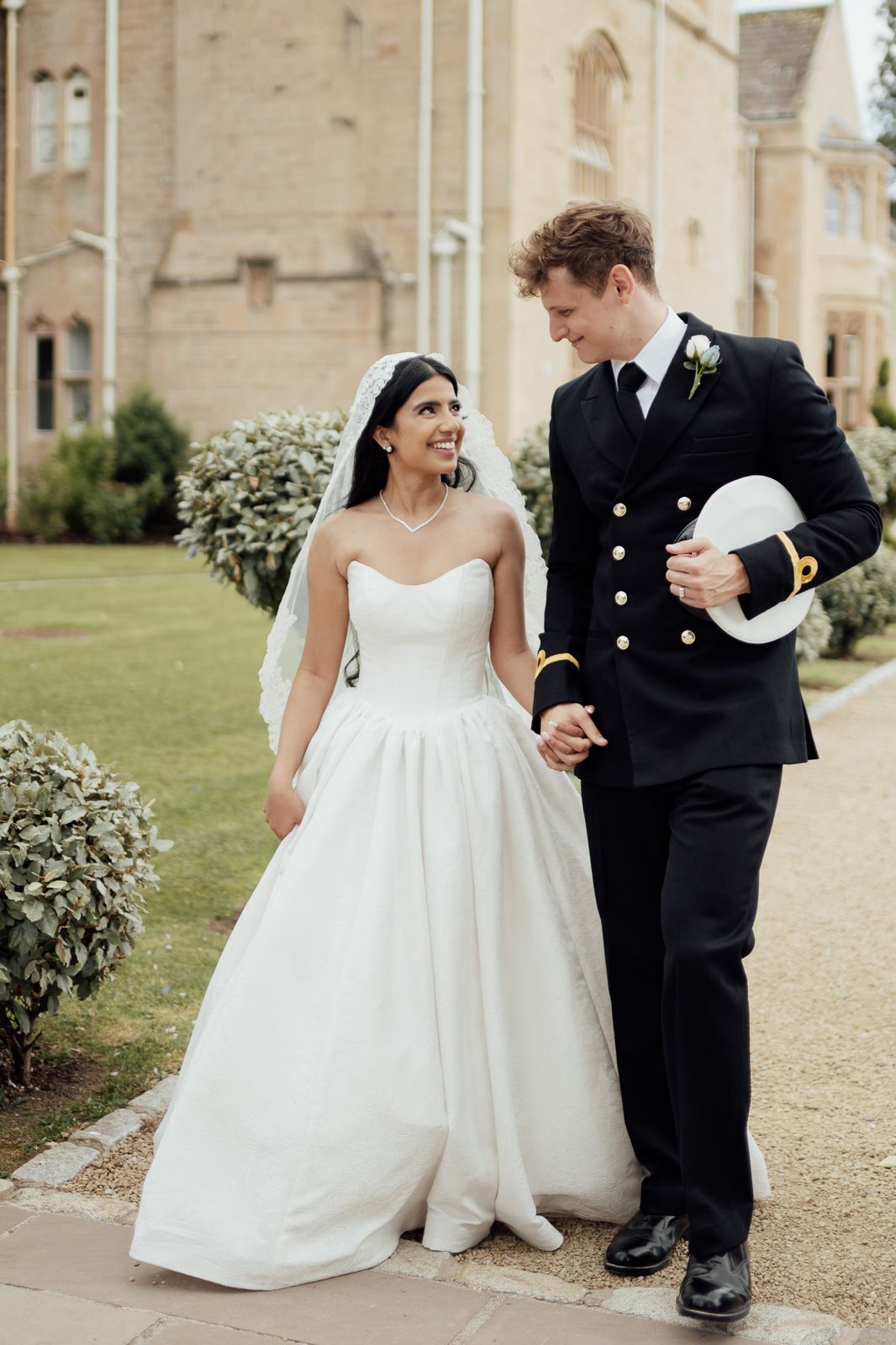 A smiling bride in a white strapless gown walks hand-in-hand with a naval officer groom outside a historic building.