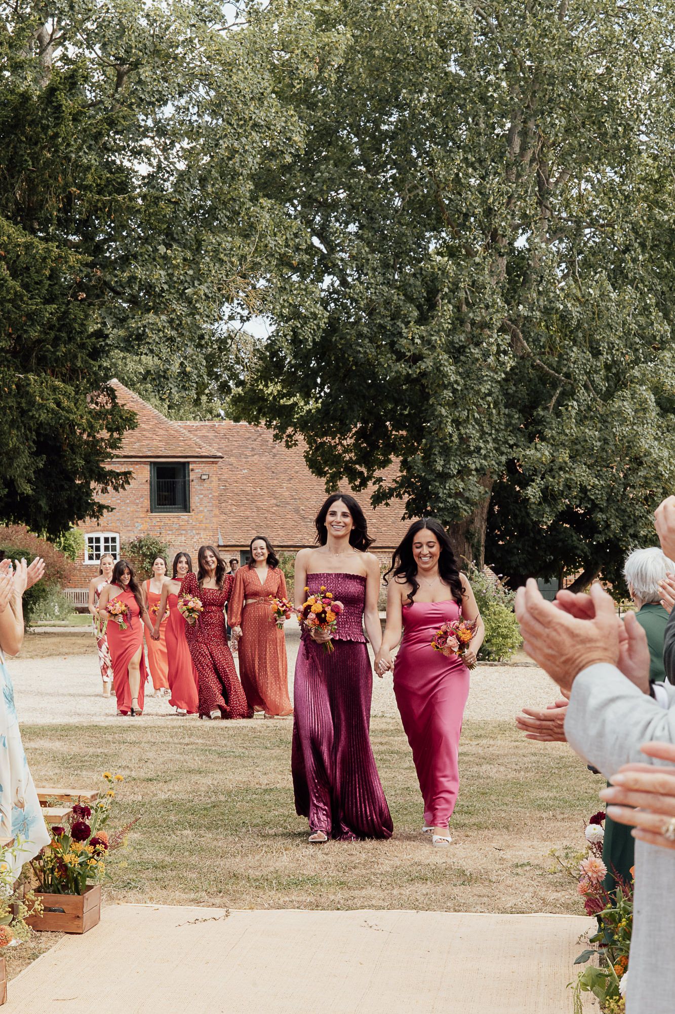 Bridesmaids in mismatched pink and red gowns walk down an outdoor aisle at a summer wedding ceremony.