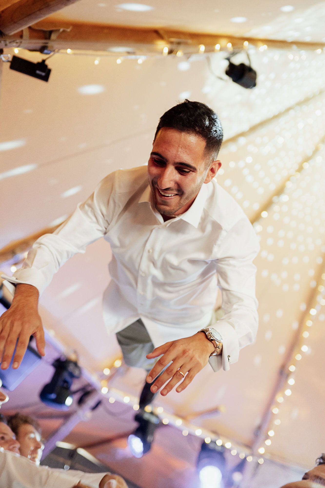 Man in white shirt smiling and dancing at a wedding reception with fairy lights in the background.