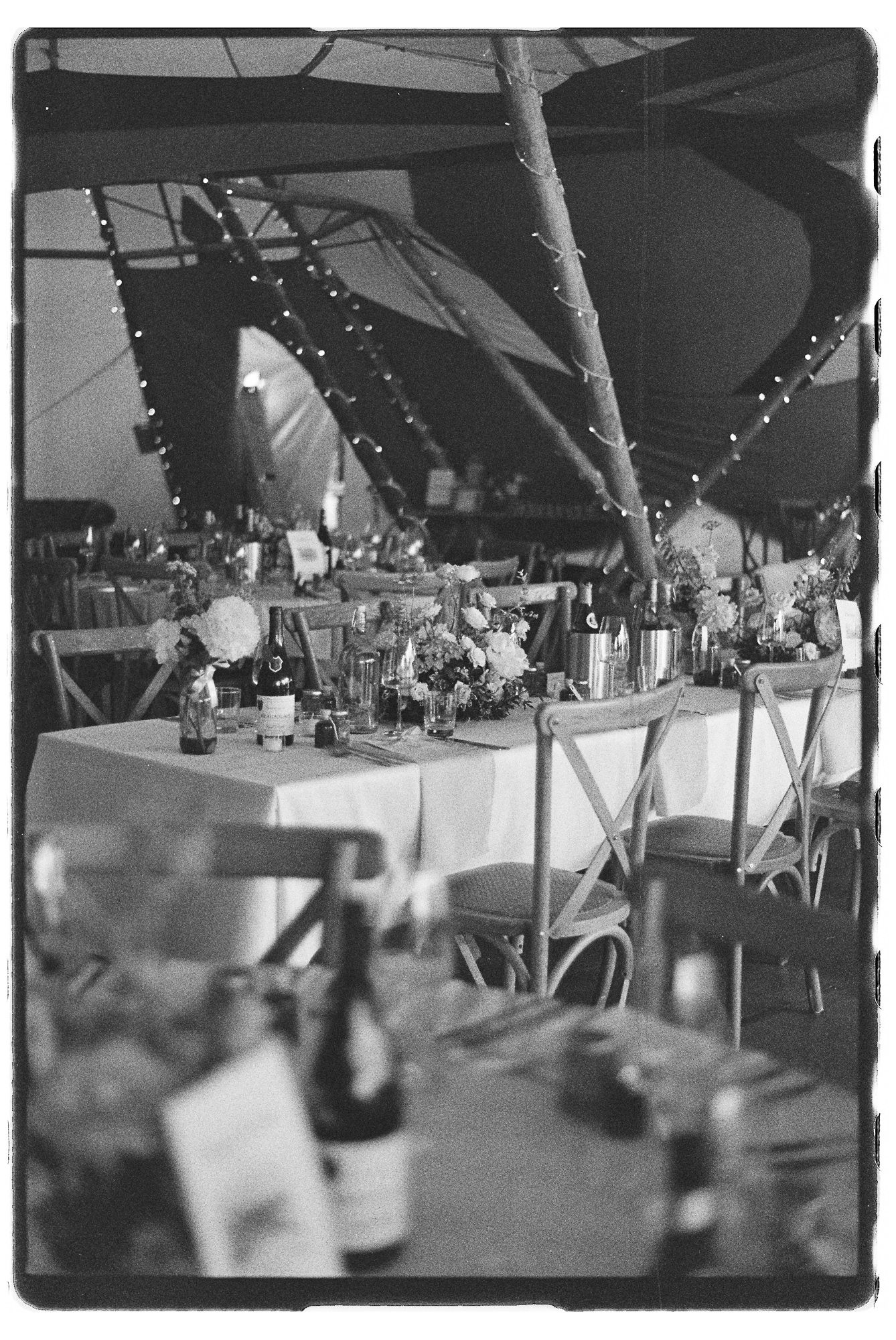 Black and white photo of an elegant outdoor wedding reception under a tent with string lights and floral centerpieces.