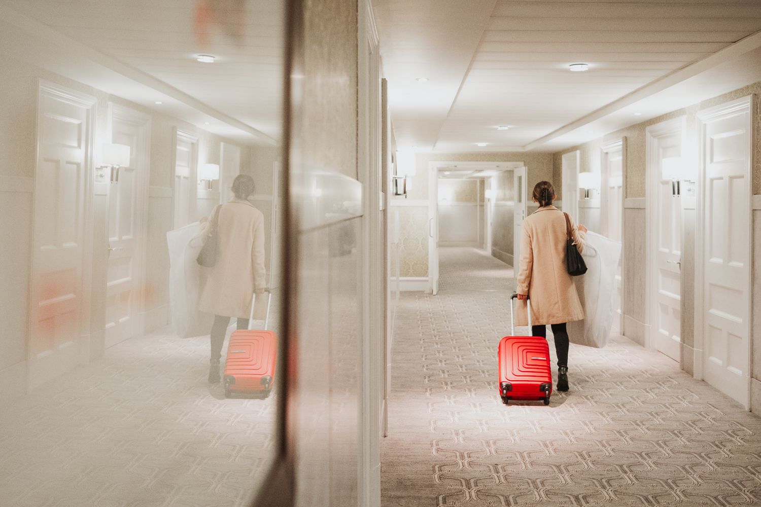 Person in beige coat walking down hotel hallway with red suitcase reflected in mirror.