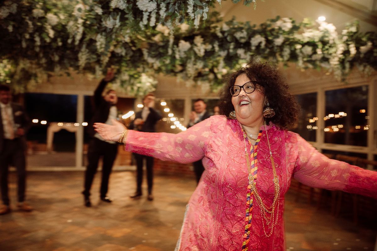 A person in a bright pink outfit dances joyfully at an indoor celebration with string lights overhead.