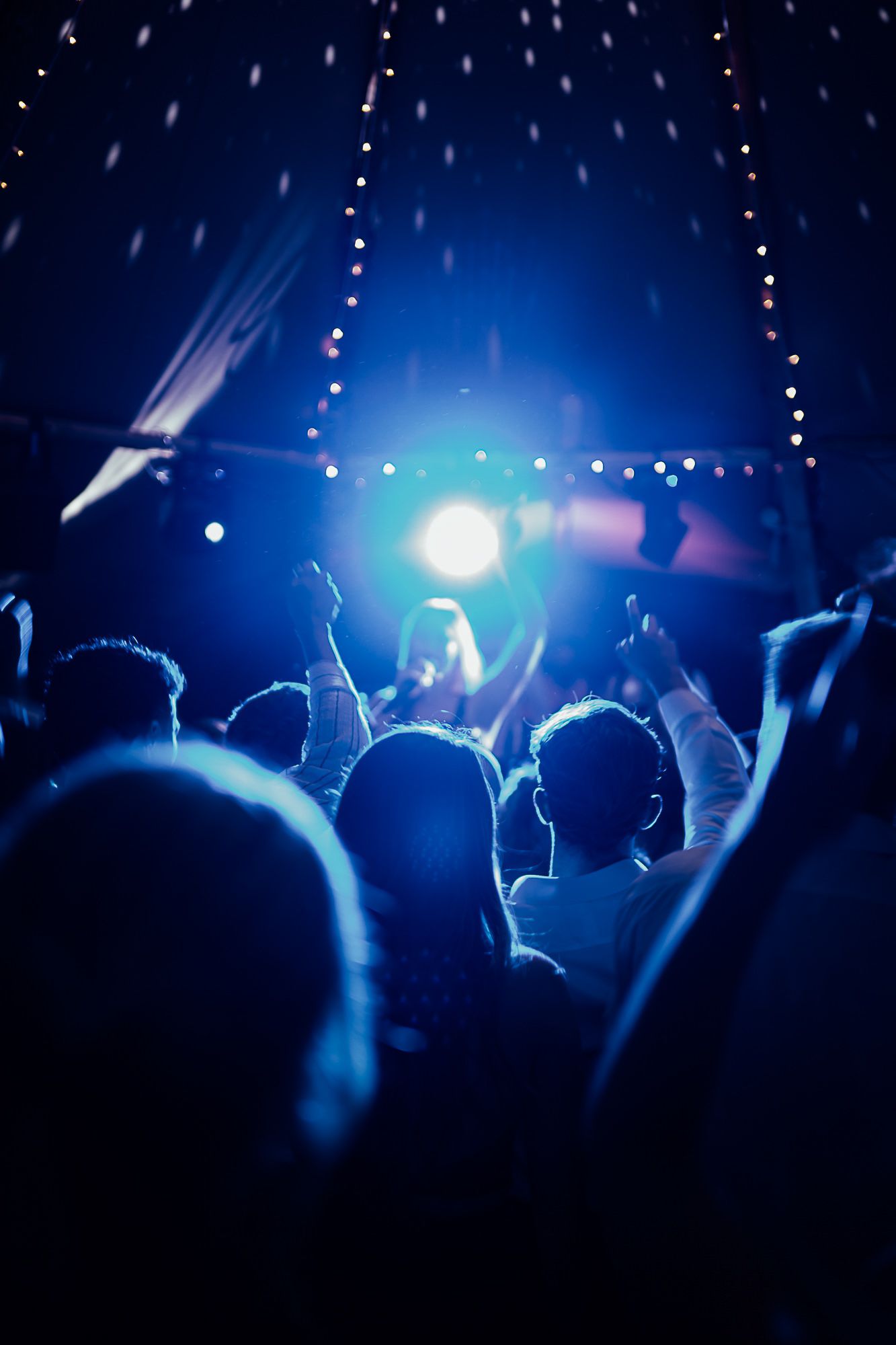 Crowd of concert-goers with raised hands under blue stage lights and bokeh string lights at a live music event.