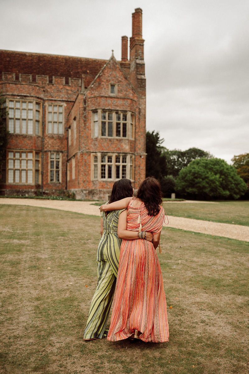 Two people in colorful dresses walk arm in arm across a lawn with a historic brick mansion in the background.