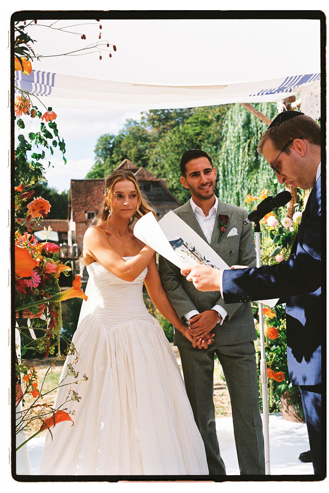 Bride and groom stand at outdoor wedding altar as officiant reads ceremony script, surrounded by colorful floral arrangements.