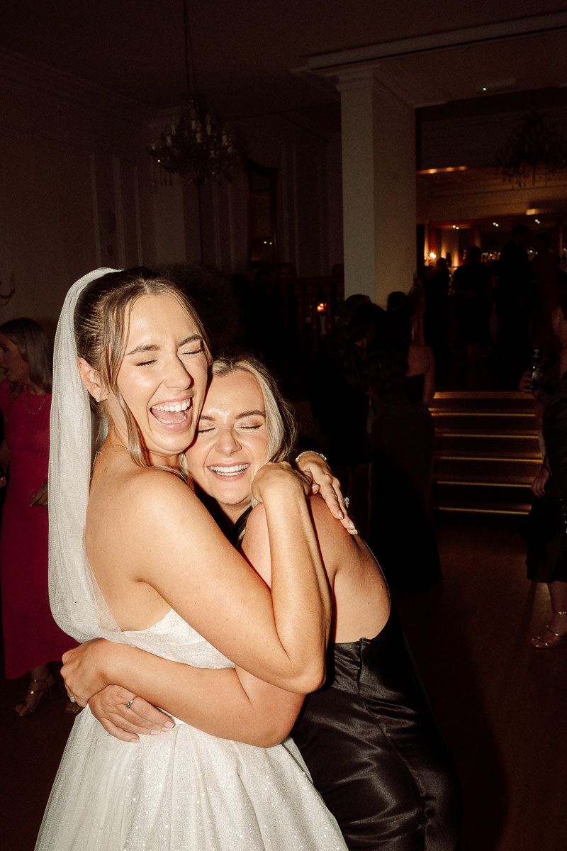 Two people share an emotional embrace while laughing joyfully at a dimly lit wedding reception.
