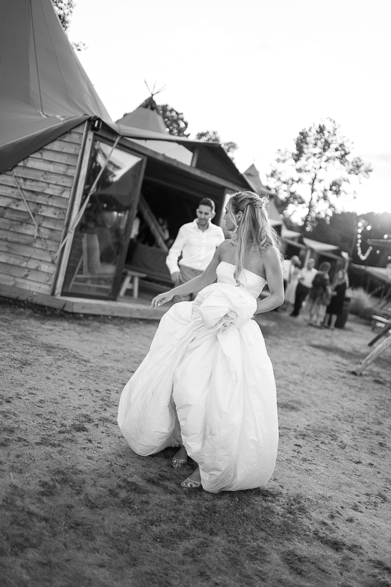 Black and white photo of a bride in a white gown dancing barefoot outdoors near a tipi tent at a wedding reception.