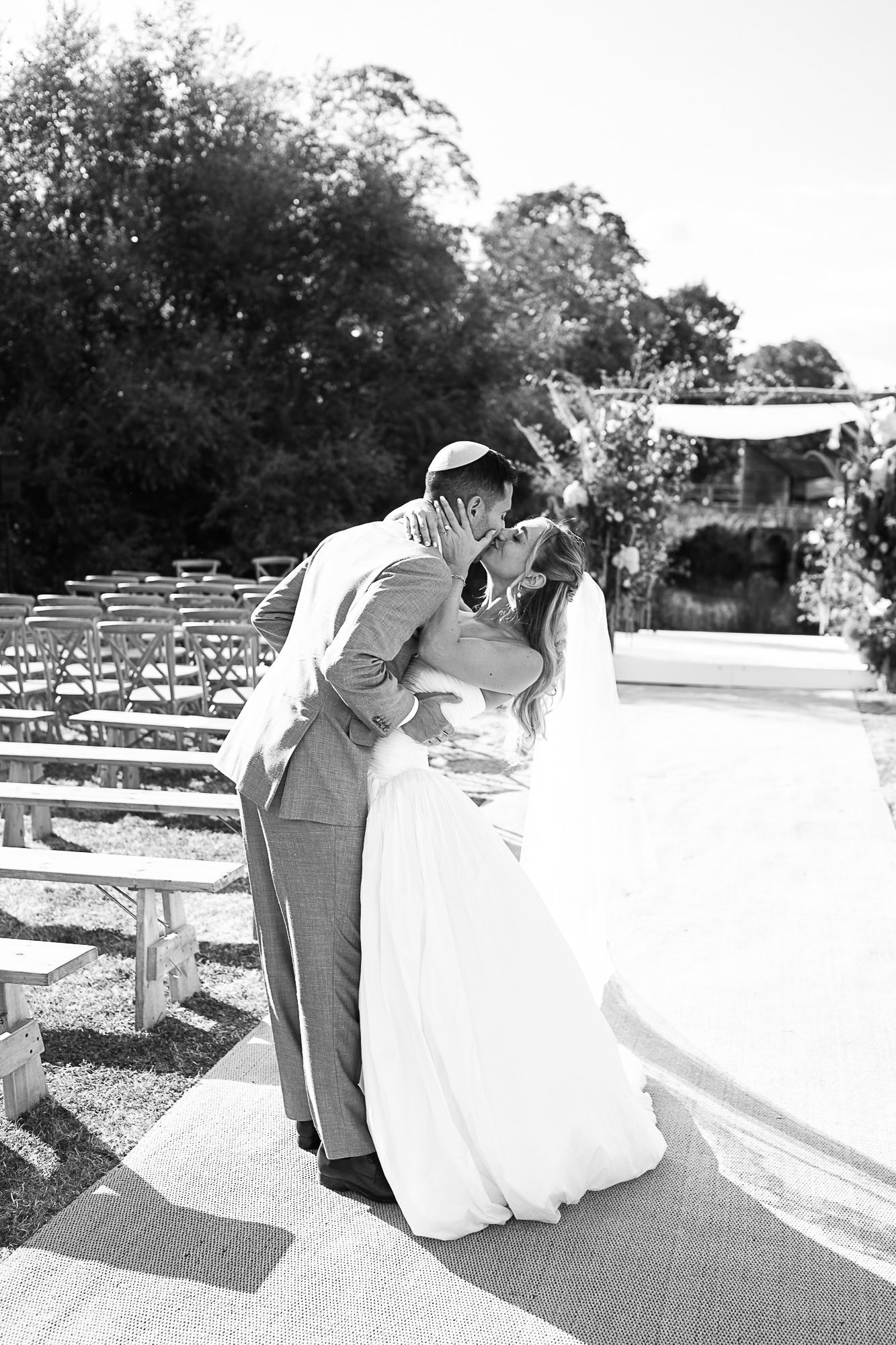 Black and white photo of bride and groom sharing first kiss at outdoor wedding ceremony with rows of chairs.