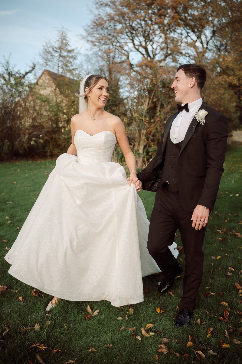 A newlywed couple playfully poses on grass with autumn leaves while the bride holds her white dress.