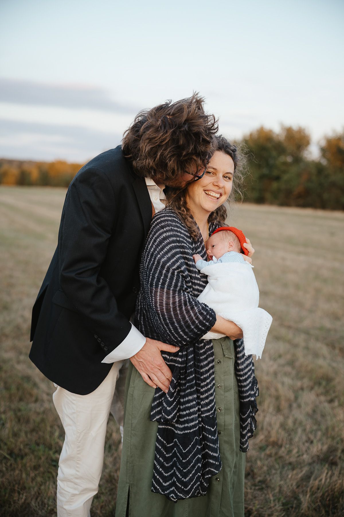 An intimate family portrait in a fall field shows a couple embracing while holding their newborn baby at sunset.
