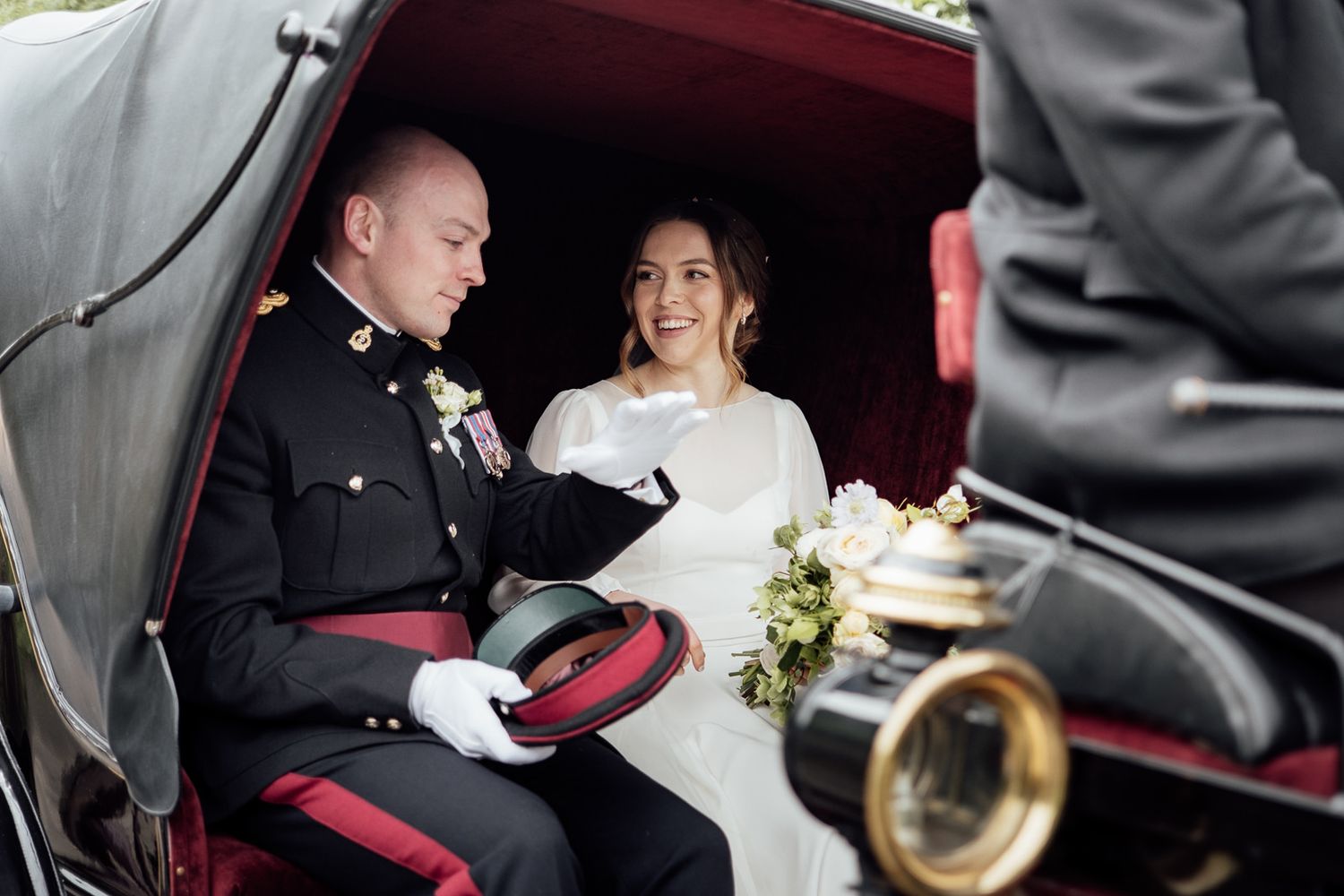 A newlywed couple sits together in a vintage carriage, with the groom in military dress uniform and bride in white gown.