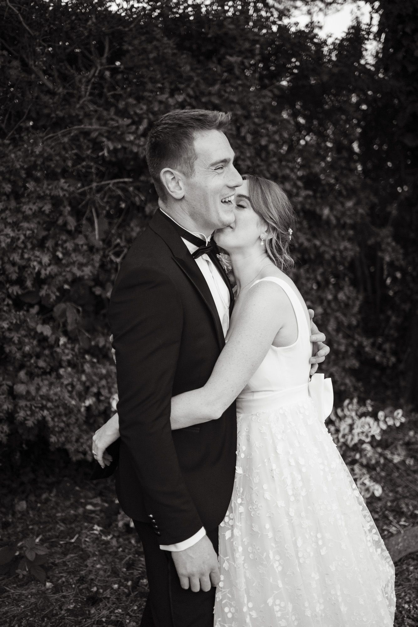 A black and white photo sequence captures an intimate kiss between newlyweds against a dark foliage background.