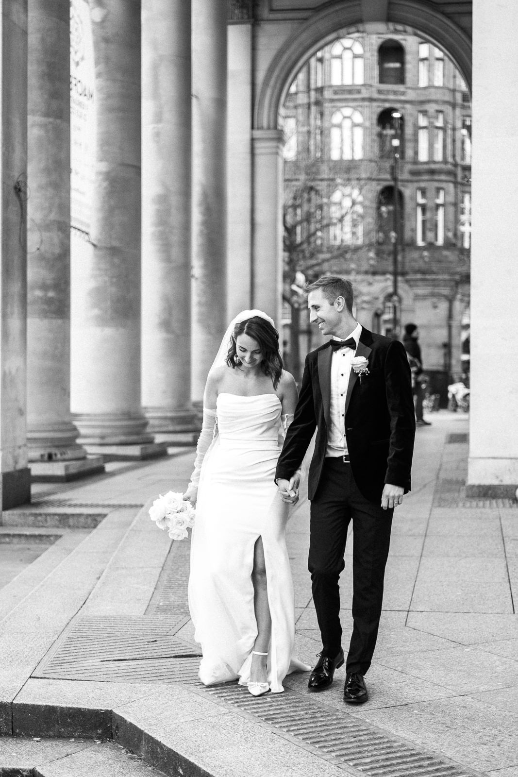 A wedding couple strolls through ornate stone columns in a grand architectural setting captured in black and white.