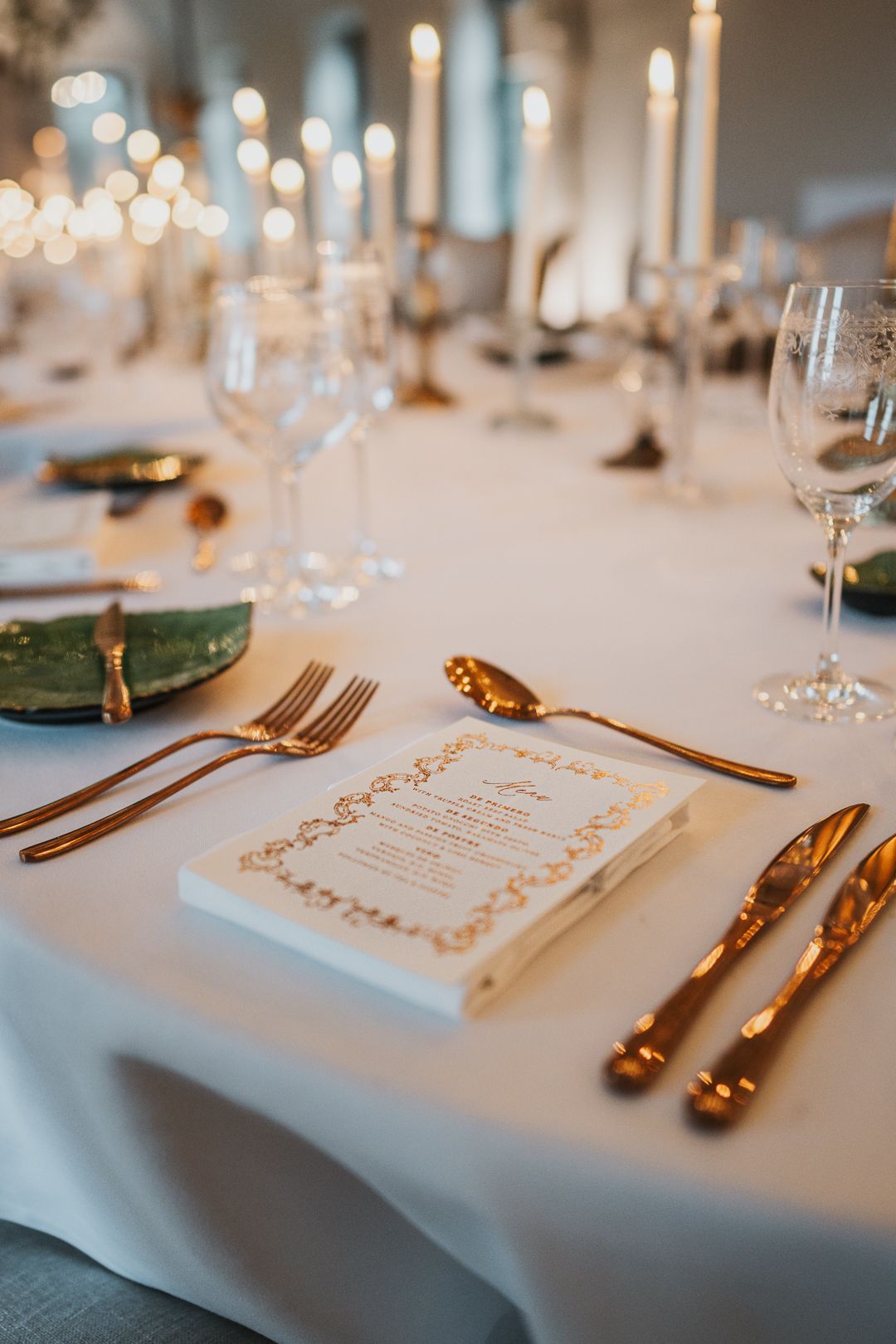 Close-up of formal place setting with gold utensils, menu card and white tablecloth at wedding reception.