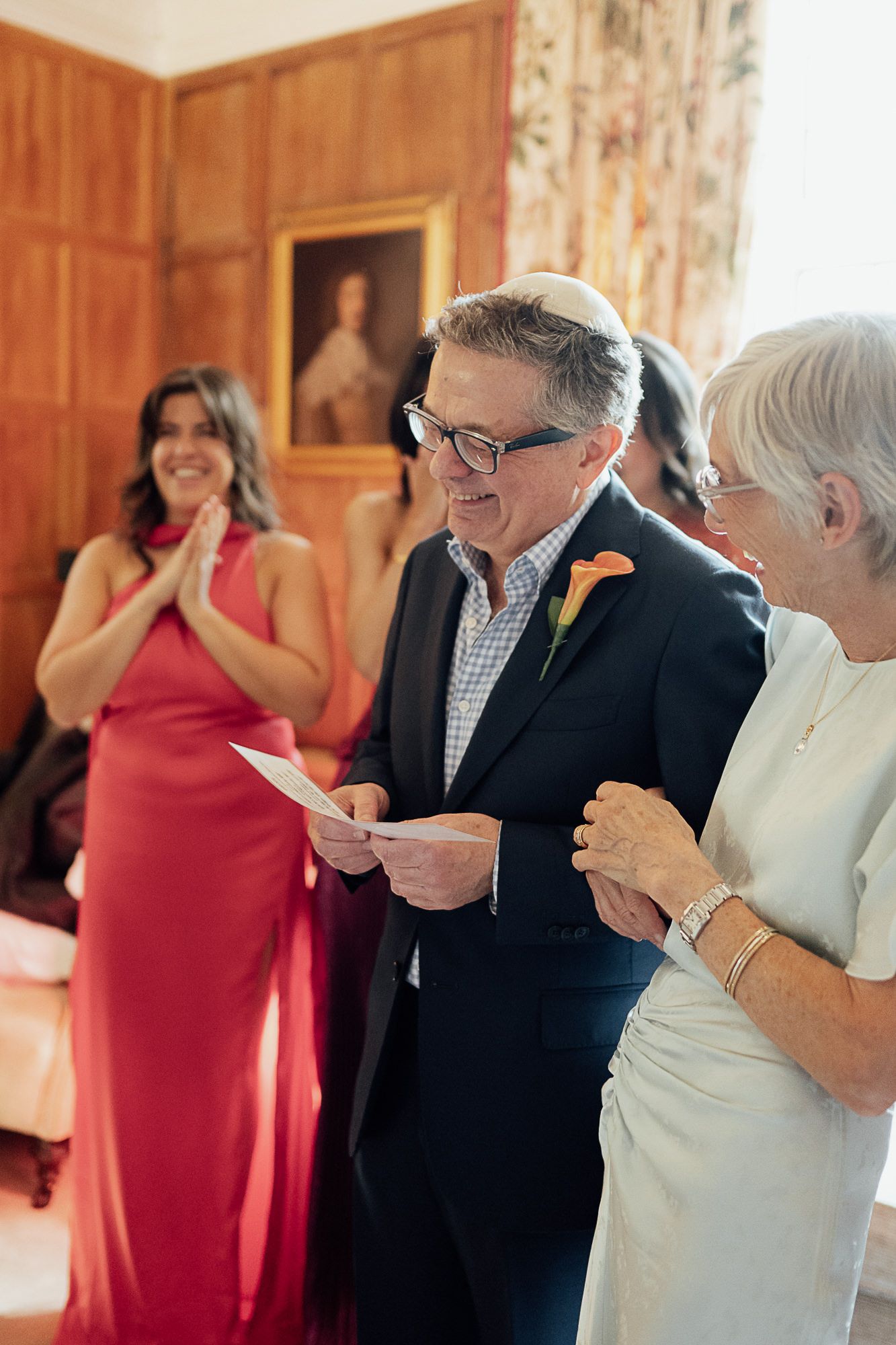 Elderly couple sharing a joyful moment at a wedding ceremony, man in navy suit with yarmulke reading a card.