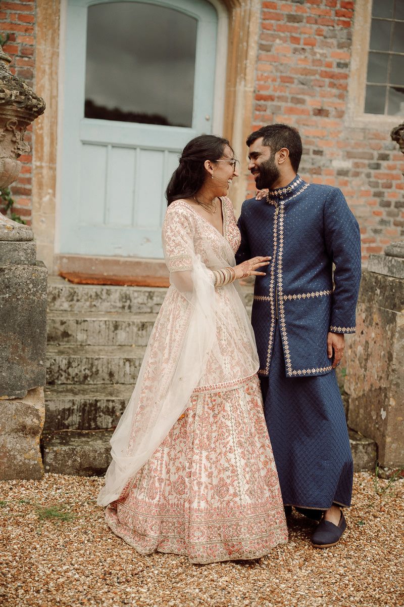 A couple in traditional South Asian attire, with the woman in pink lehenga and man in blue kurta, pose together outdoors at mapledurham.
