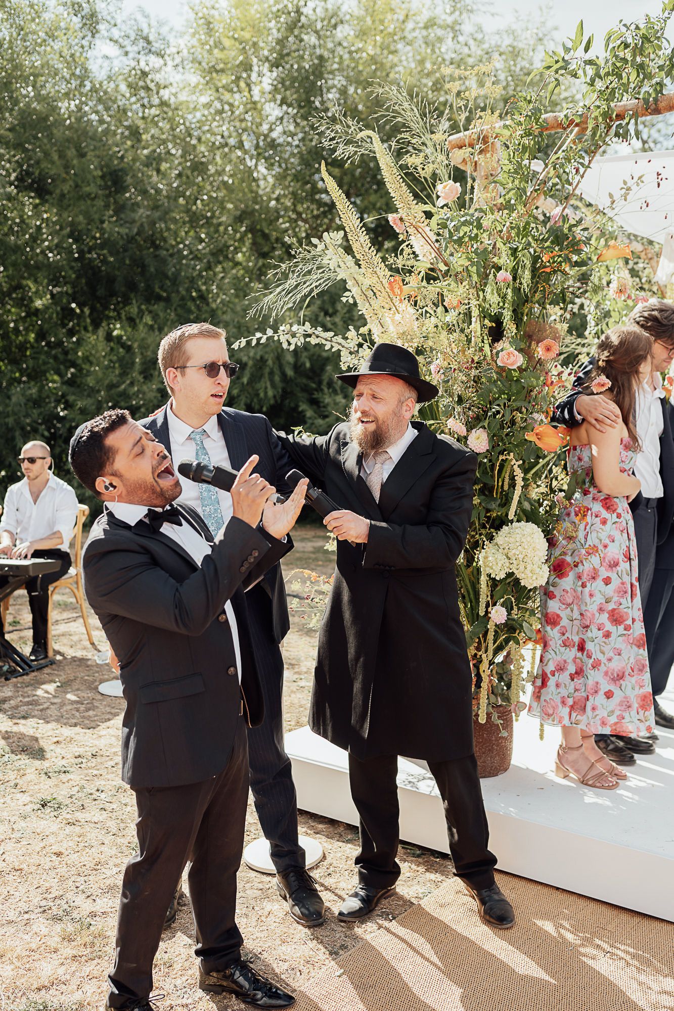 Men in formal attire laugh and celebrate outdoors at a Jewish wedding ceremony near a floral chuppah.