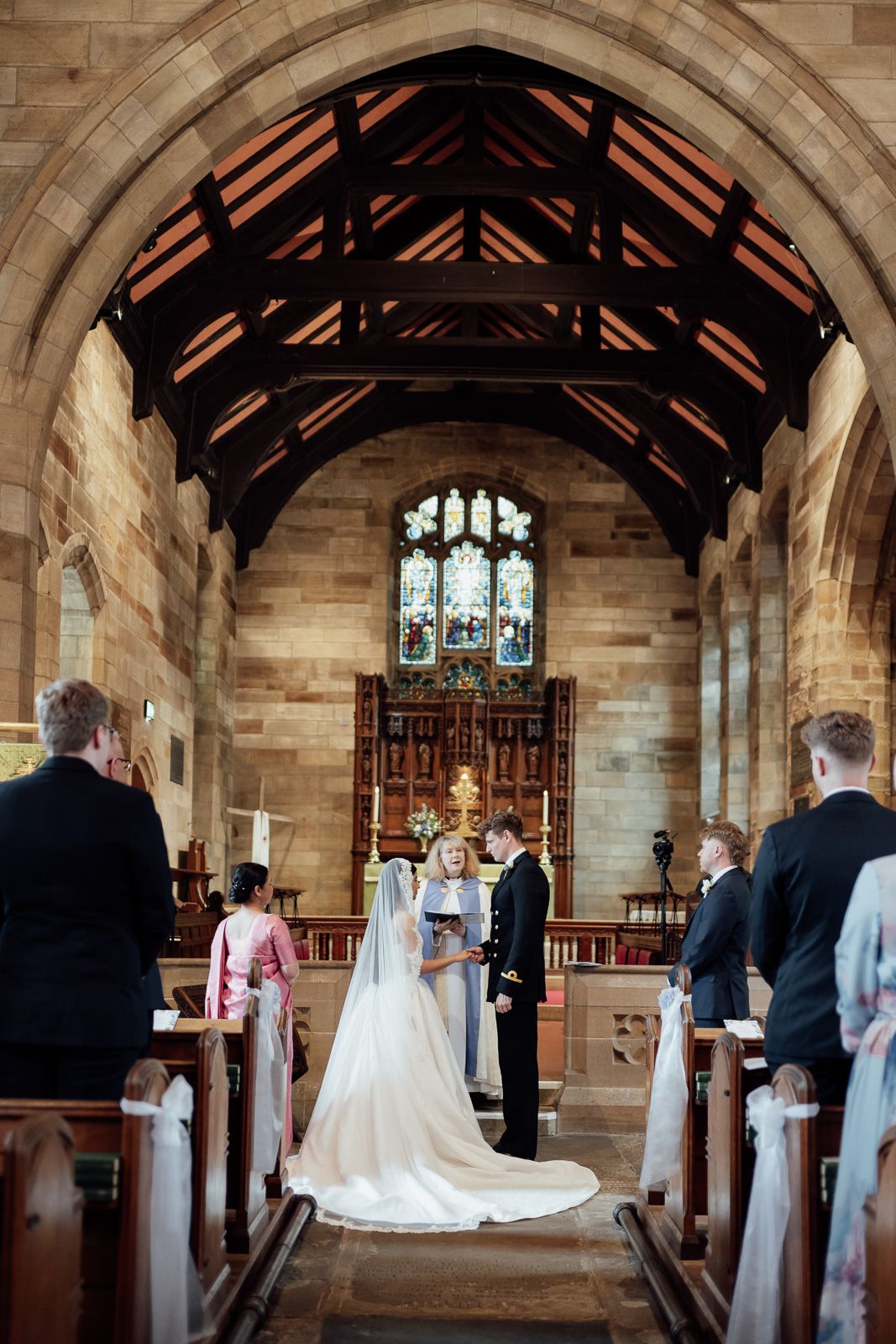Bride and groom exchange vows at the altar of a traditional stone church with wooden beamed ceiling and stained glass window.