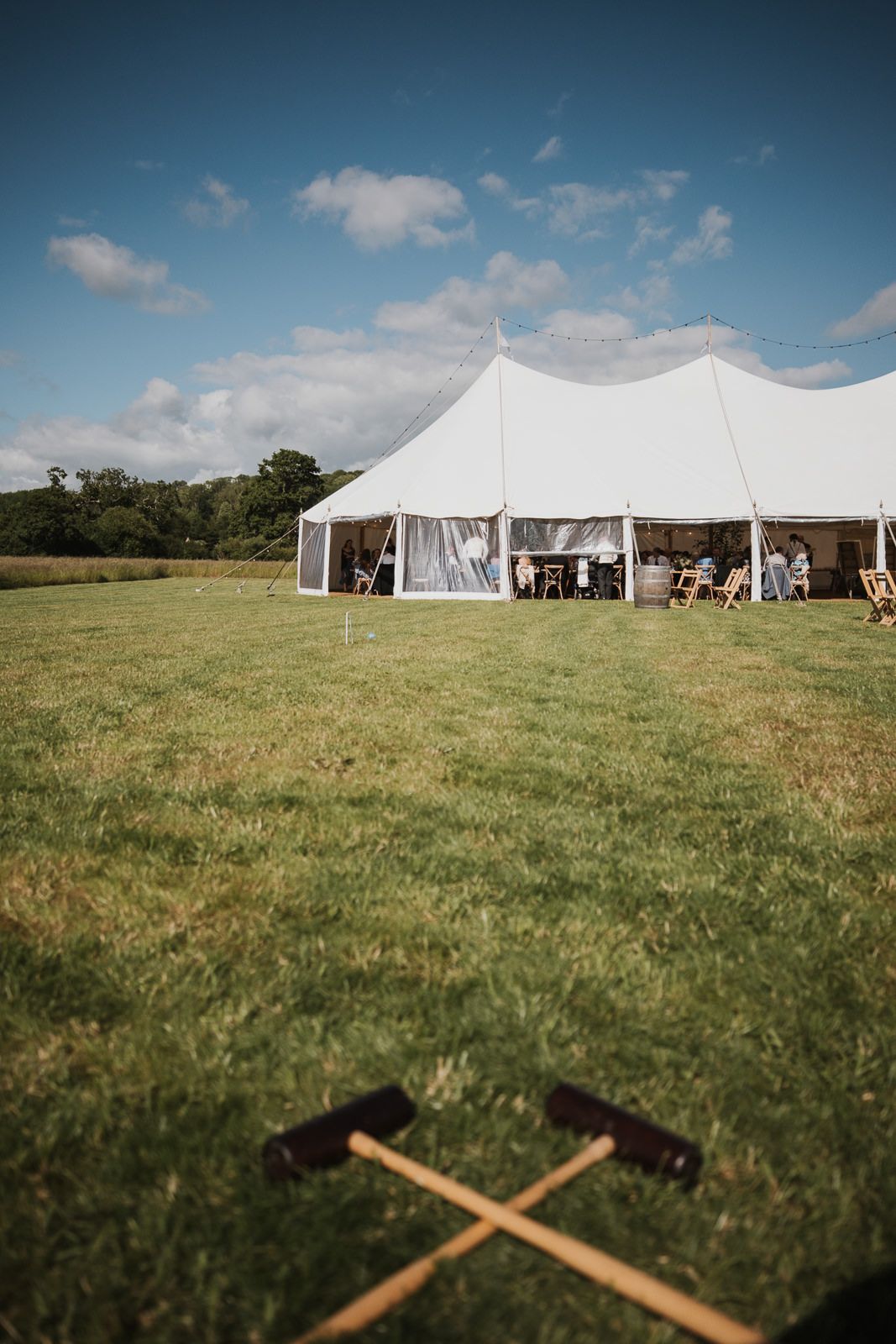 Large white wedding tent set up in a grassy field with croquet mallets crossed in the foreground under blue summer sky.